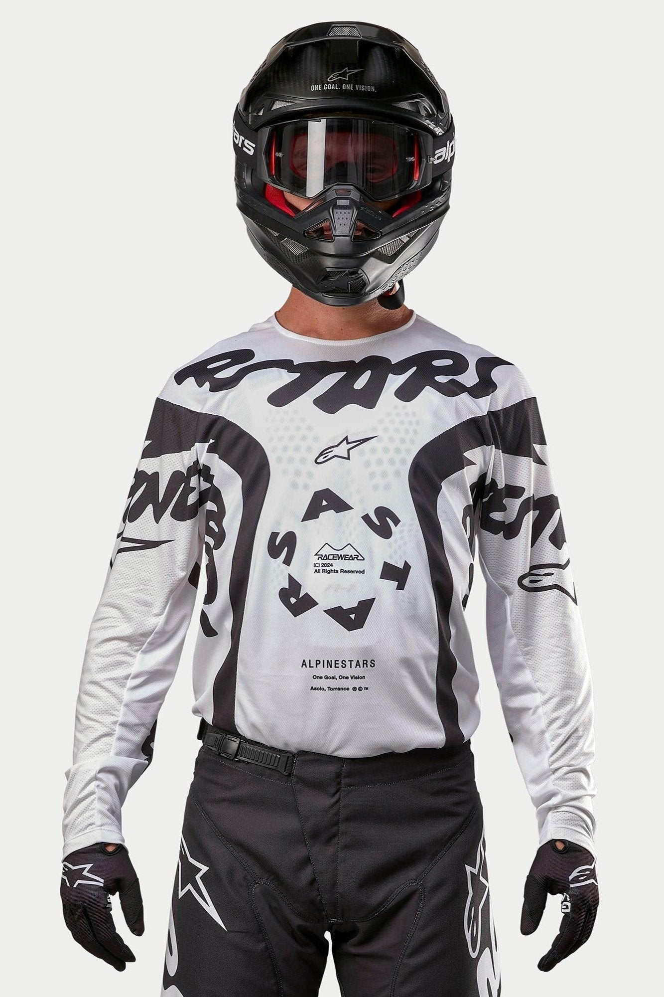 Zawodnik motocrossowy ubrany w koszulkę Alpinestars 2024 Racer Hana w kolorze biało-czarnym, w zestawie z czarnym kaskiem i czarnymi spodniami motocrossowymi, z wyraźną czarną grafiką „ASTARS” i okrągłym logo na środku, profesjonalna odzież do wyścigów terenowych
