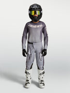Alpinestars koszulka i zestaw Fluid Amber młodzieżowe 2027, szaro-piaskowy strój motocrossowy, czarny kask i białe buty.