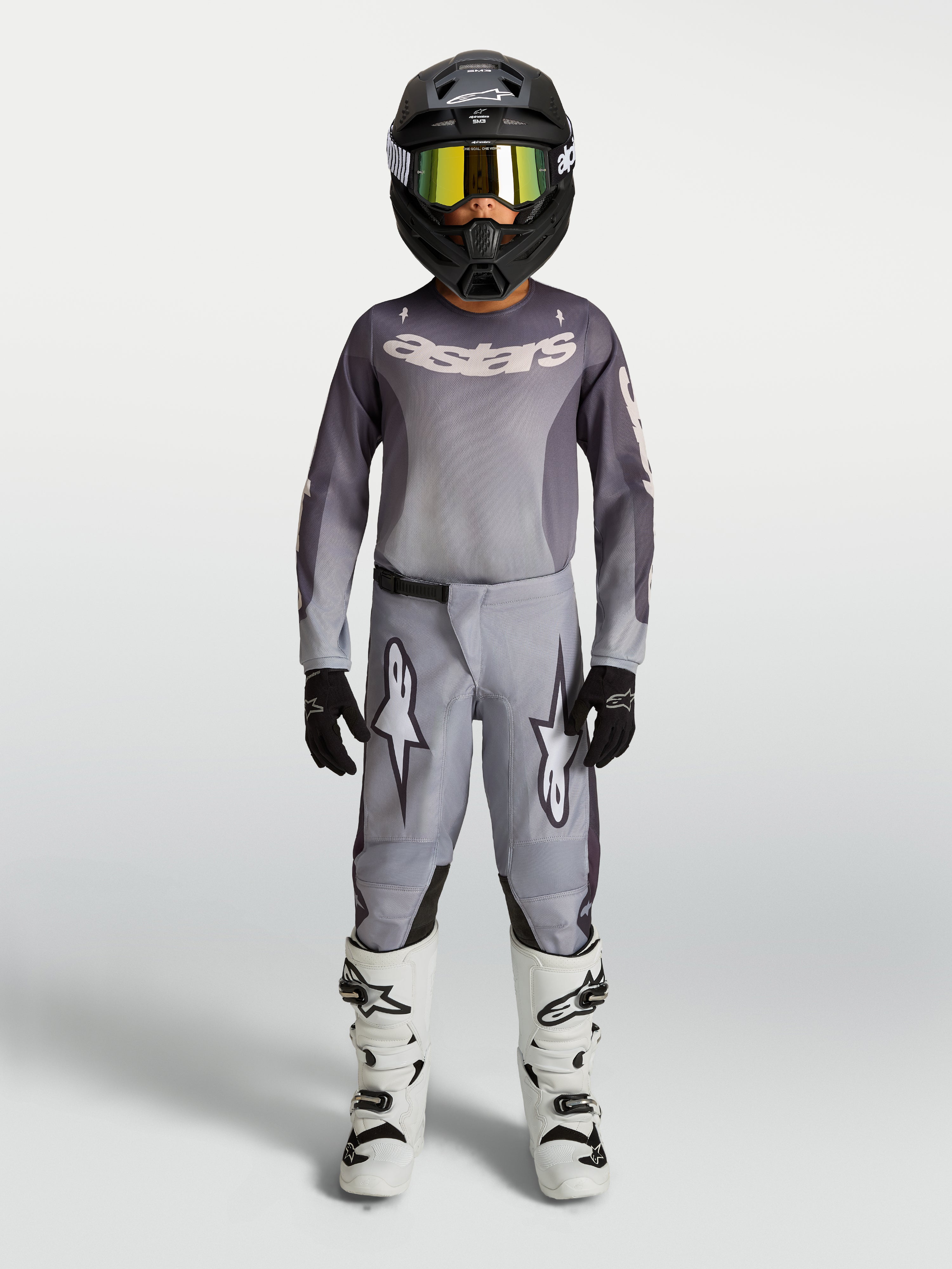 Alpinestars koszulka i zestaw Fluid Amber młodzieżowe 2027, szaro-piaskowy strój motocrossowy, czarny kask i białe buty.
