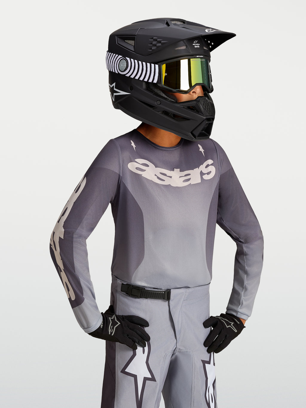 Alpinestars Koszulka Fluid Amber Młodzieżowe 2027, piaskowo-szara koszulka motocrossowa na modelu w czarnym kasku wyścigowym.