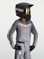 Alpinestars Koszulka Fluid Amber Młodzieżowe 2027, piaskowo-szara koszulka motocrossowa na modelu w czarnym kasku wyścigowym.