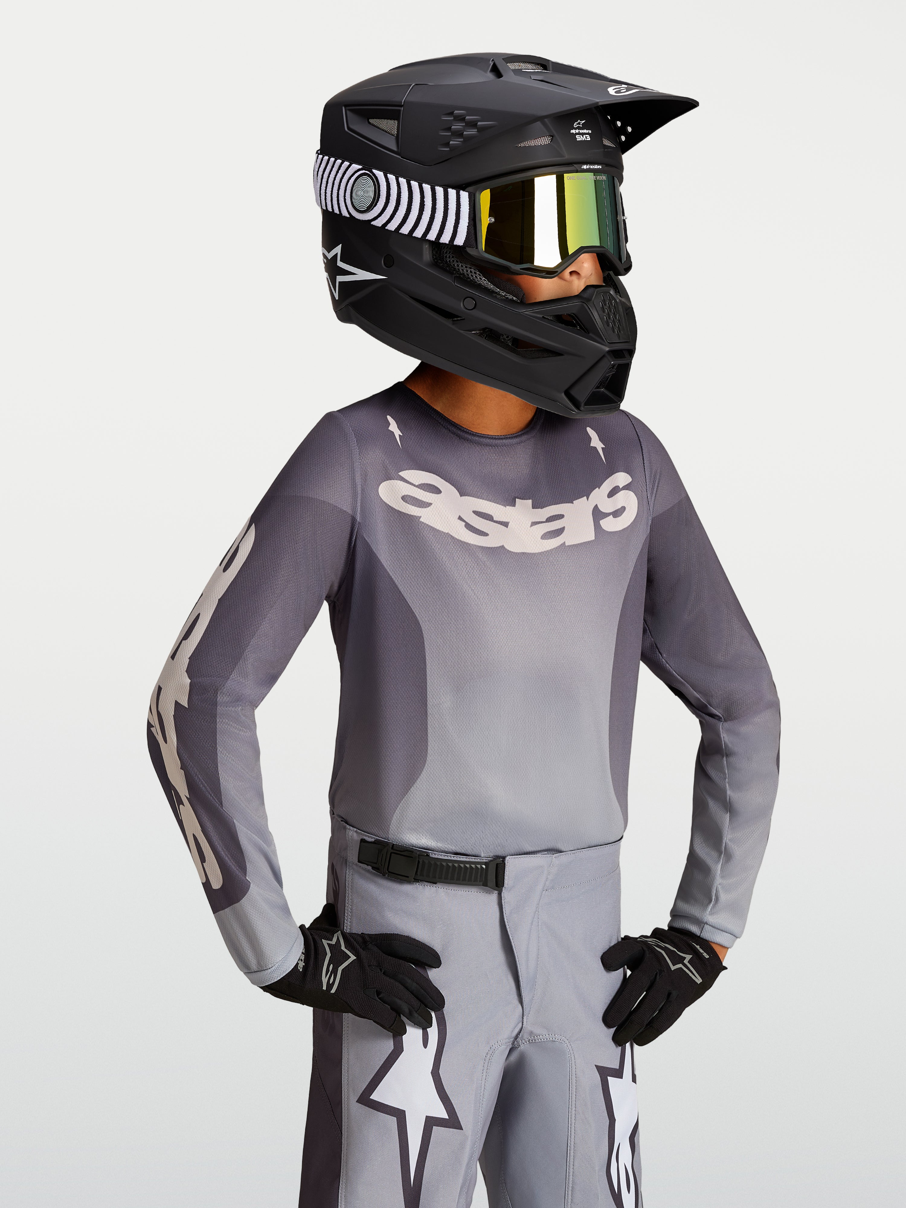 Alpinestars Koszulka Fluid Amber Młodzieżowe 2027, piaskowo-szara koszulka motocrossowa na modelu w czarnym kasku wyścigowym.