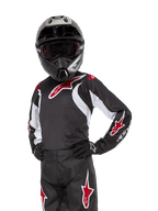 Alpinestars Youth 2024 Racer Lucent Jersey, młodzieżowa koszulka motocrossowa, czarno-biało-czerwona, noszona przez młodego zawodnika w czarnym kasku i pasujących spodniach, z czerwonymi logotypami gwiazdy i białymi akcentami graficznymi, wyczynowa odzież off-road