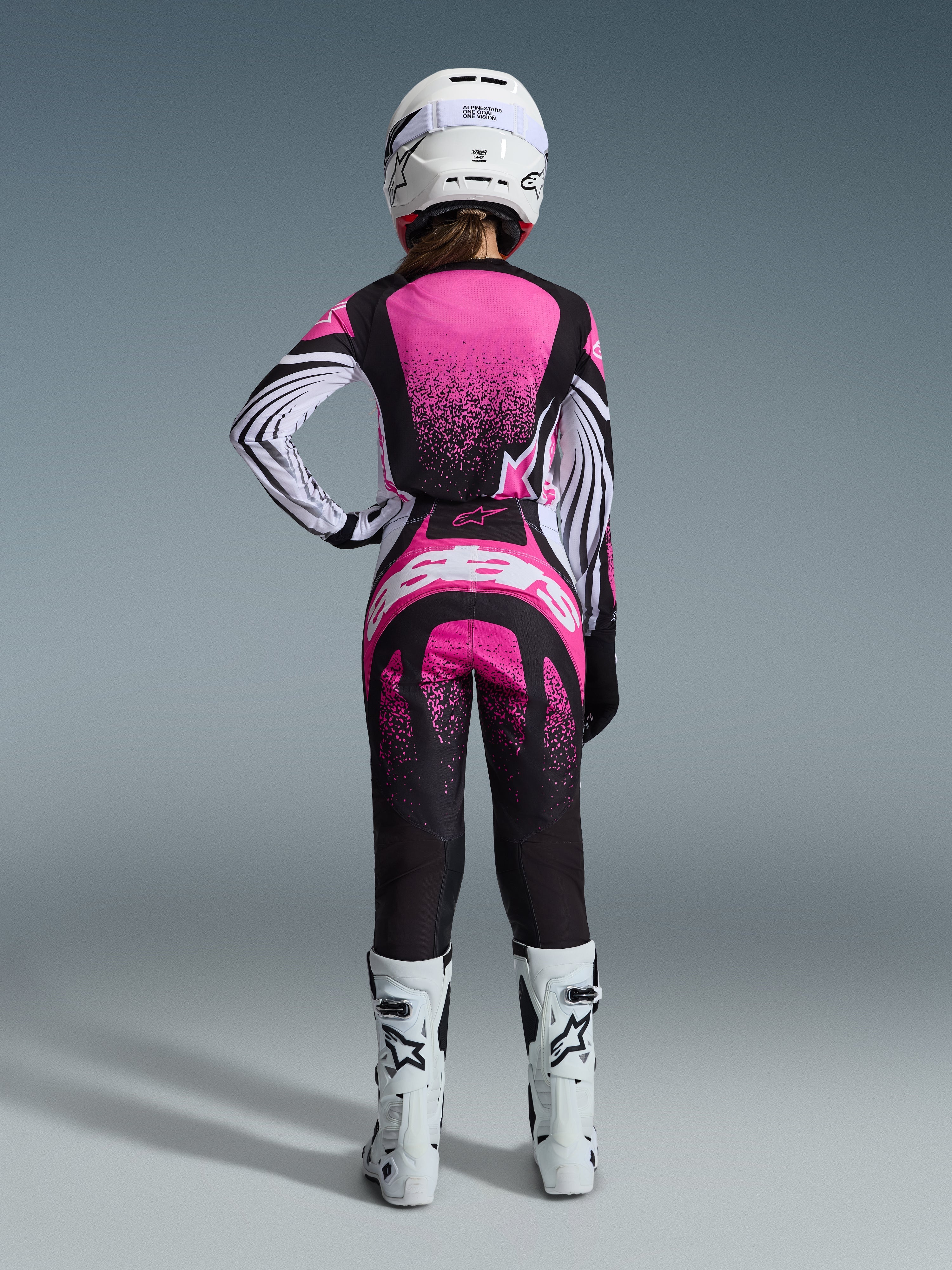 Alpinestars Stella Techstar Nomur Gear Set 2026, zawodniczka motocrossowa widoczna od tyłu, różowo-czarna koszulka z nakrapianym wzorem i pasiastymi rękawami, pasujące spodnie z białym logo, biały kask i białe buty off-road