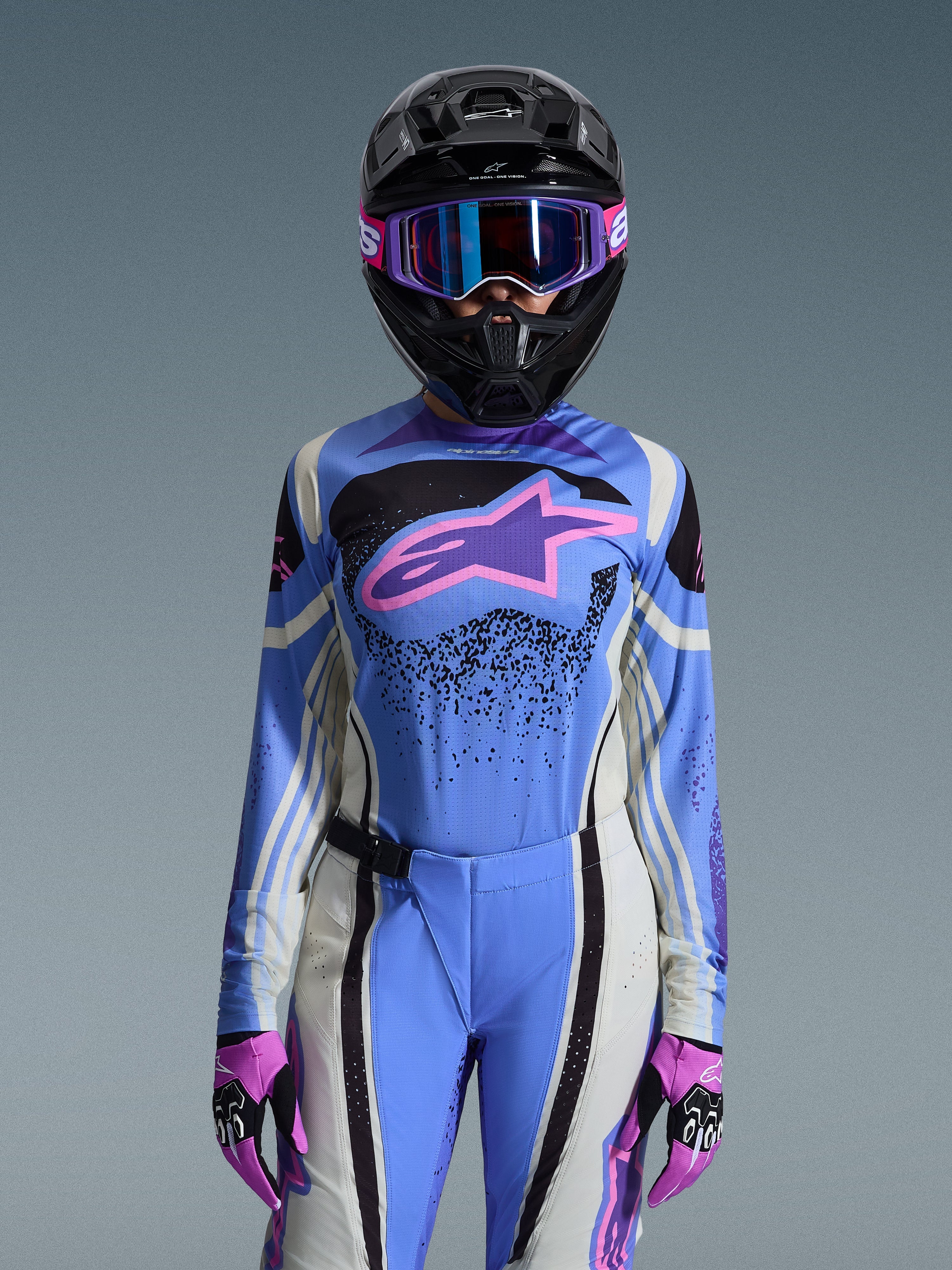 Zawodniczka motocrossu w zestawie Alpinestars 2026 Stella Techstar Nomur, niebieska koszulka z różowo-fioletowym logo gwiazdy, pasujące niebieskie spodnie z biało-czarnymi panelami bocznymi, czarny kask z fioletowo-niebieskimi goglami i różowo-czarne rękawice, kompletny damski strój wyścigowy off-road