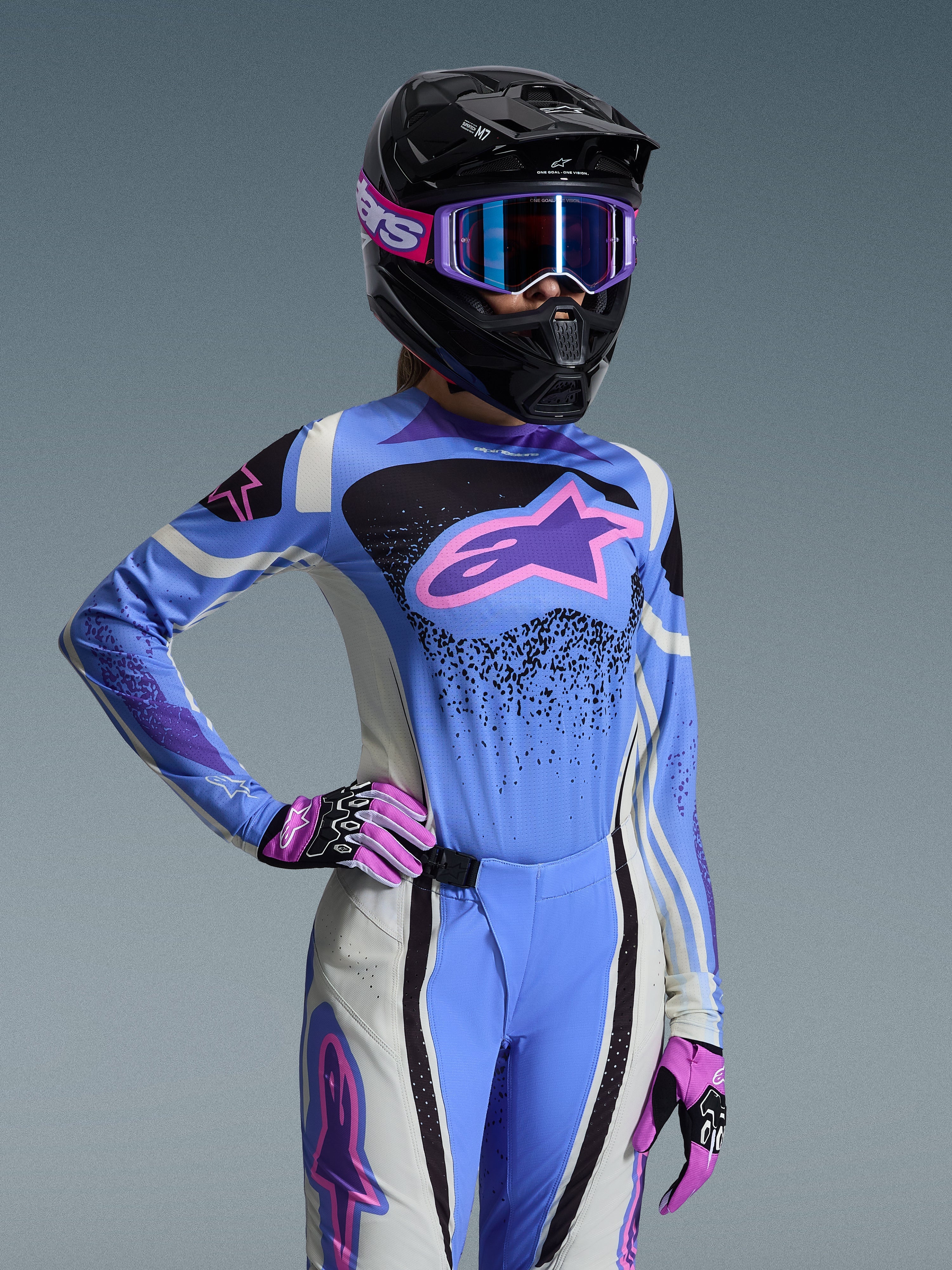Zawodniczka motocrossu w zestawie Alpinestars 2026 Stella Techstar Nomur, jasnoniebieska koszulka z różowymi i fioletowymi akcentami, pasujące niebiesko-białe spodnie, czarny kask z fioletowymi goglami, różowo-czarne rękawice, profesjonalny strój off-road dla kobiet.