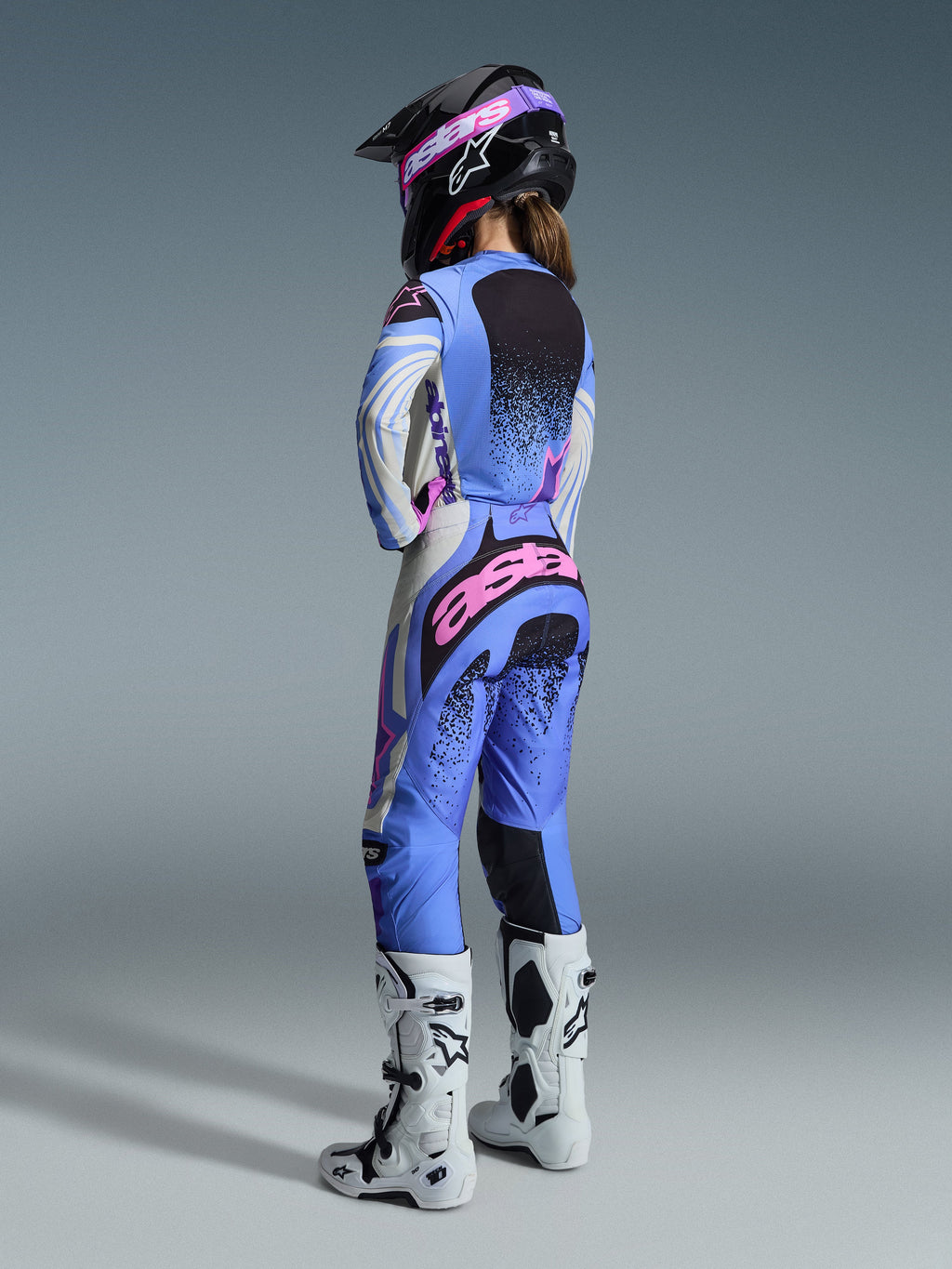 Alpinestars 2026 Stella Techstar Nomur Gear Set, widok z tyłu na zawodniczkę motocrossową w jasnoniebieskim, białym i różowym stroju z czarnymi nakrapianymi detalami, czarny kask i białe buty, profesjonalny zestaw wyścigowy dla kobiet
