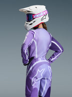 Zawodniczka motocrossowa w zestawie Alpinestars 2026 Stella Fluid Apex, fioletowa i lawendowa koszulka oraz spodnie z białą grafiką, biały kask z różowo-fioletowymi goglami, dedykowany damski krój, dopasowany strój do wyścigów off-road