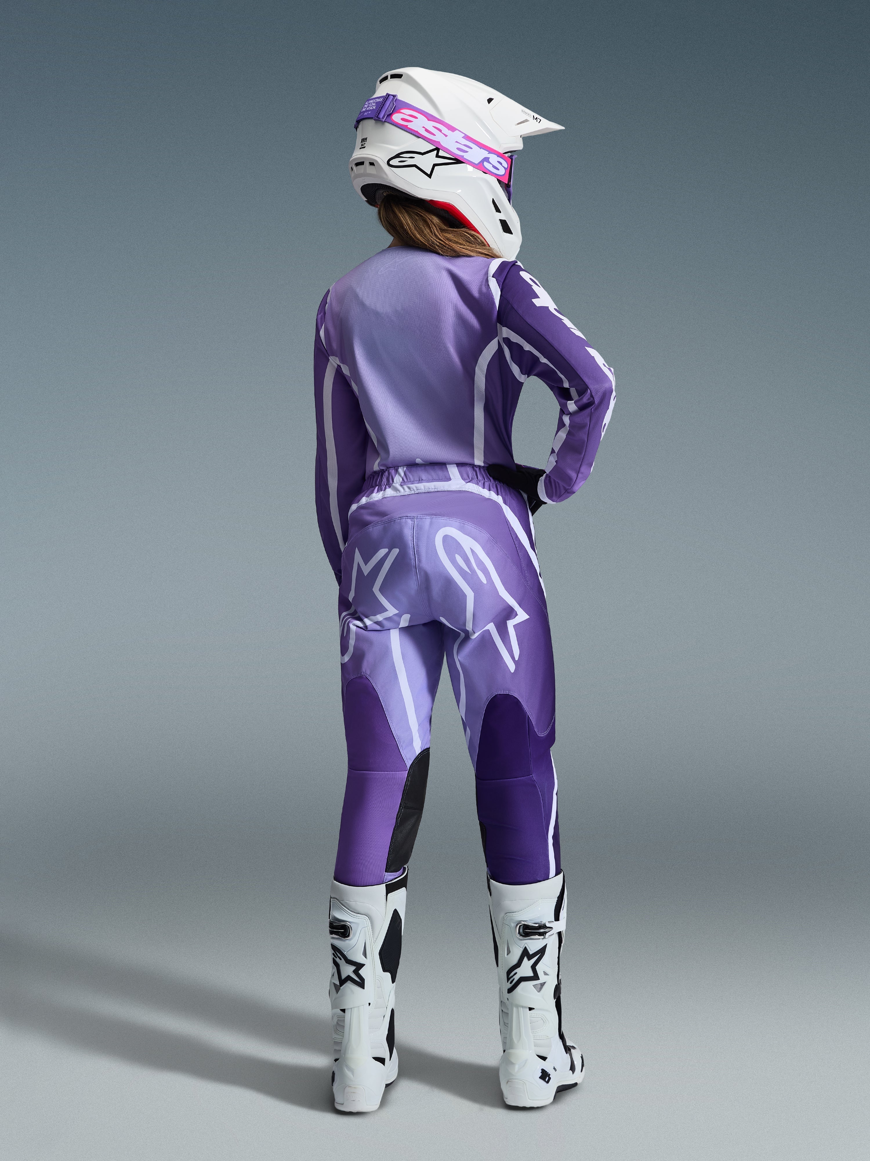 Alpinestars 2026 Stella Fluid Apex Gear Set, damska odzież motocrossowa, fioletowy i lawendowy z białymi akcentami, motocyklistka widziana od tyłu w dopasowanej koszulce i spodniach, biały kask z różowymi goglami i białe buty, skoordynowany sportowy strój wyścigowy