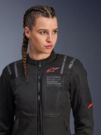 Alpinestars Stella ST-2 Air Jacket, damska kurtka motocyklowa w kolorze czarnym, z panelami z siatki zapewniającymi przepływ powietrza, brandingiem „Alpinestars Exploration Equipment” ze współrzędnymi i czerwonym logo na klatce piersiowej, prezentowana przez modelkę z warkoczami.