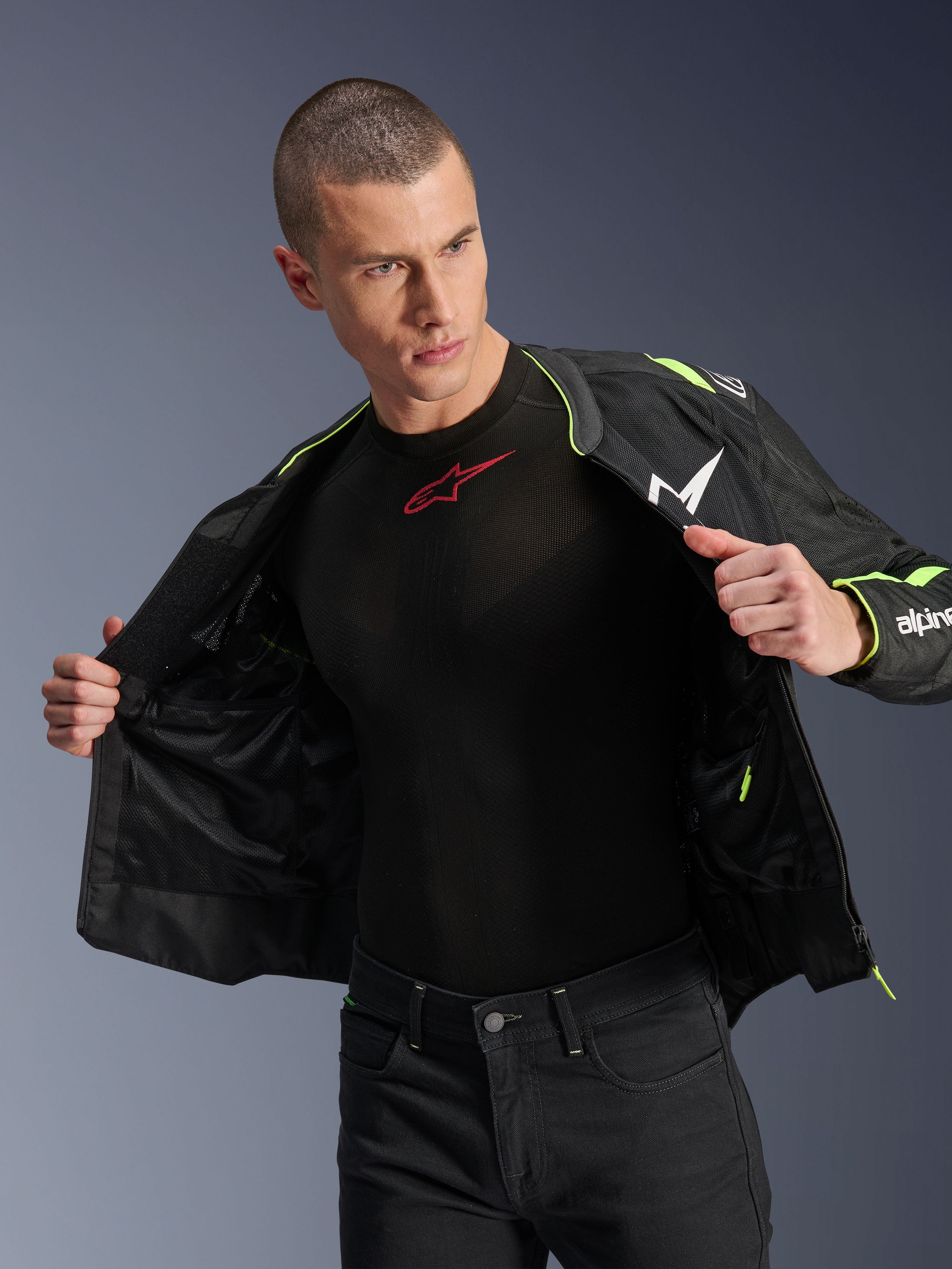 Męski model ubrany w kurtkę Alpinestars T-Sps Air V2 w kolorze czarnym i żółtym fluo, rozpiętą, aby wyeksponować wewnętrzną konstrukcję z siatki i kieszenie, w zestawieniu z czarną warstwą bazową Alpinestars i czarnymi spodniami, miejski styl odzieży motocyklowej