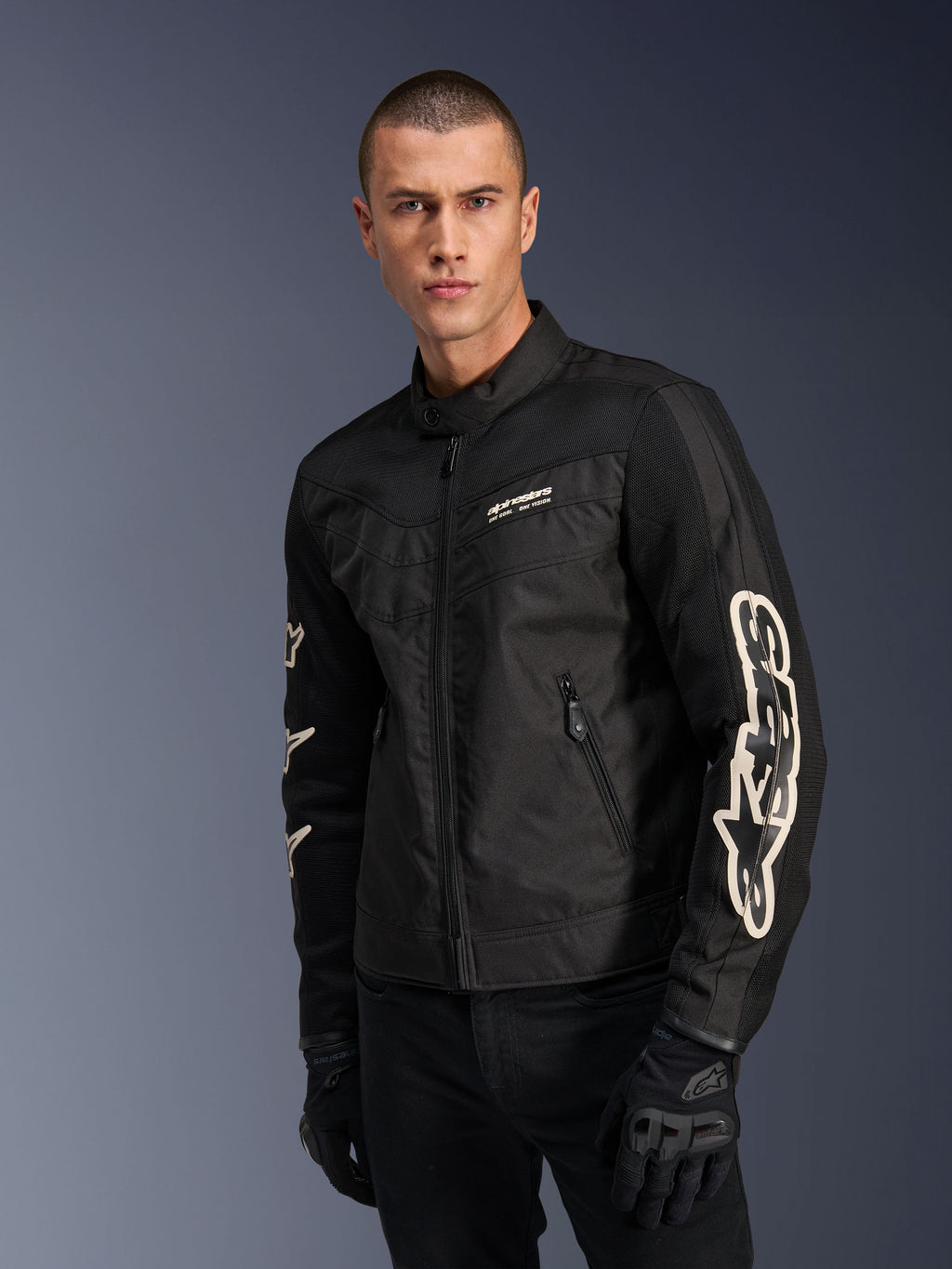 Alpinestars T-Dyno Air Jacket w kolorze czarnym, model męski w tekstylnej kurtce motocyklowej inspirowanej stylem vintage, panele z siatki zapewniające wentylację, duże białe logo Alpinestars na rękawach, czarne rękawice motocyklowe, miejski design odzieży motocyklowej
