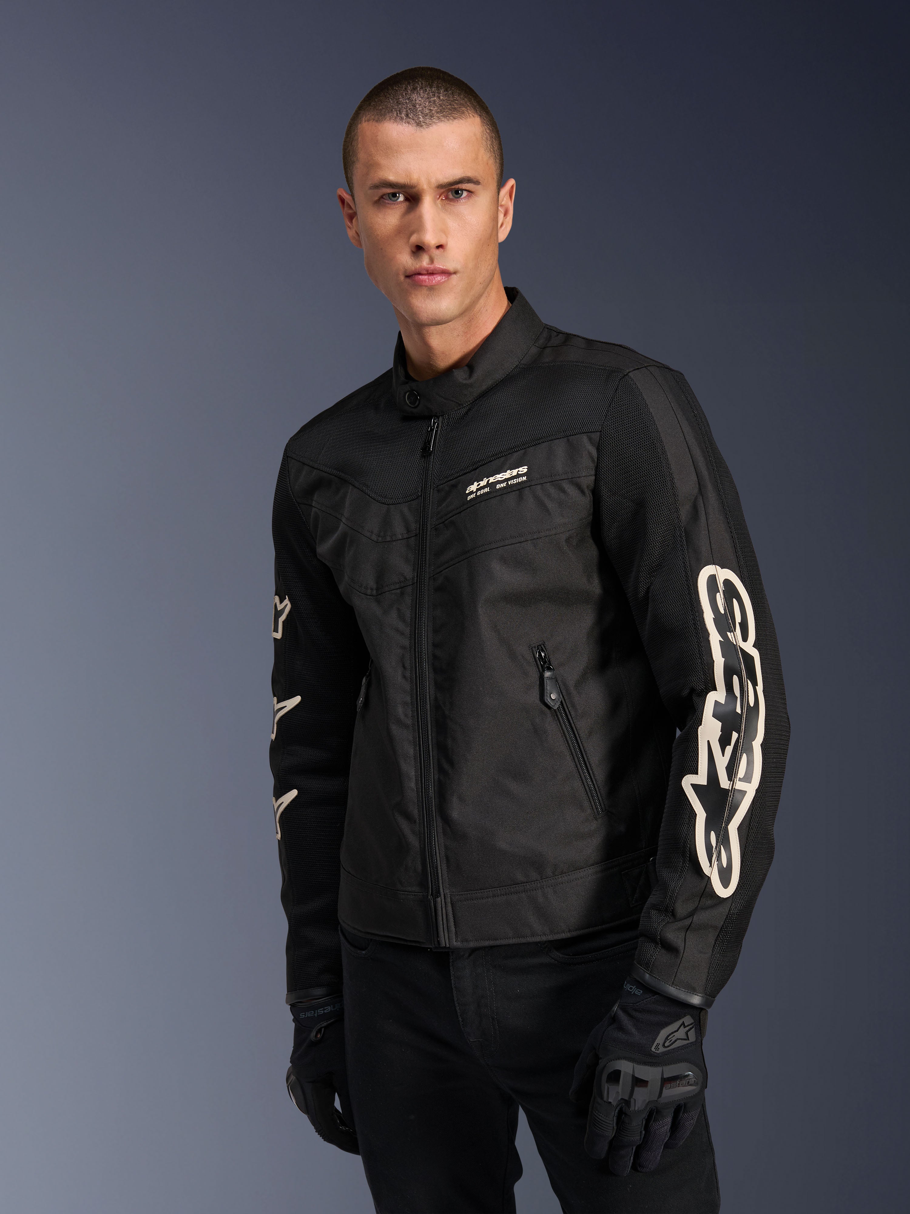 Alpinestars T-Dyno Air Jacket w kolorze czarnym, model męski w tekstylnej kurtce motocyklowej inspirowanej stylem vintage, panele z siatki zapewniające wentylację, duże białe logo Alpinestars na rękawach, czarne rękawice motocyklowe, miejski design odzieży motocyklowej