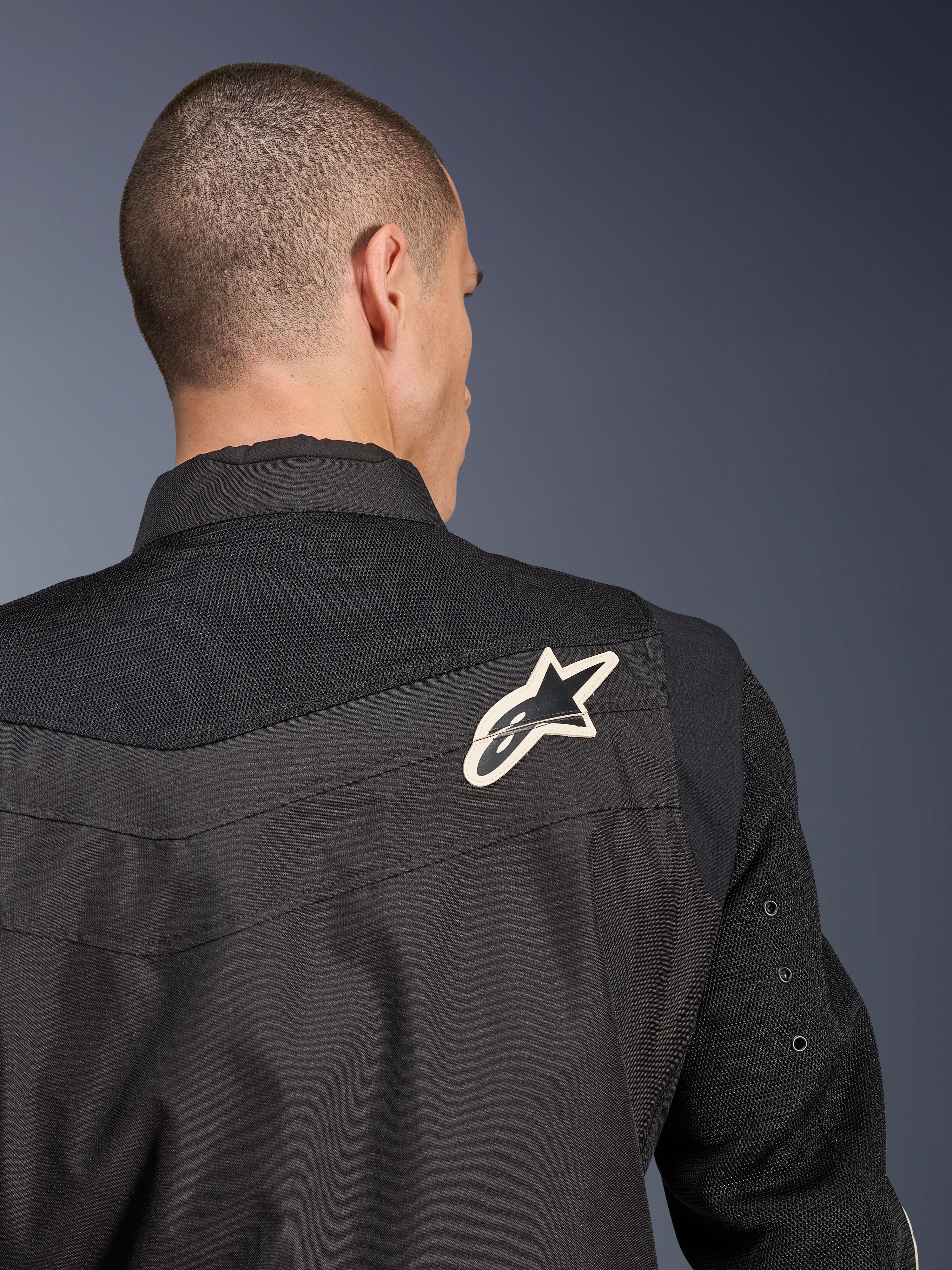 Alpinestars T-Dyno Air Jacket, czarna tekstylna kurtka motocyklowa, zbliżenie na tył podkreślające oddychający karczek i rękawy z siatki 3D, z biało-czarnym logo gwiazdy na prawym ramieniu