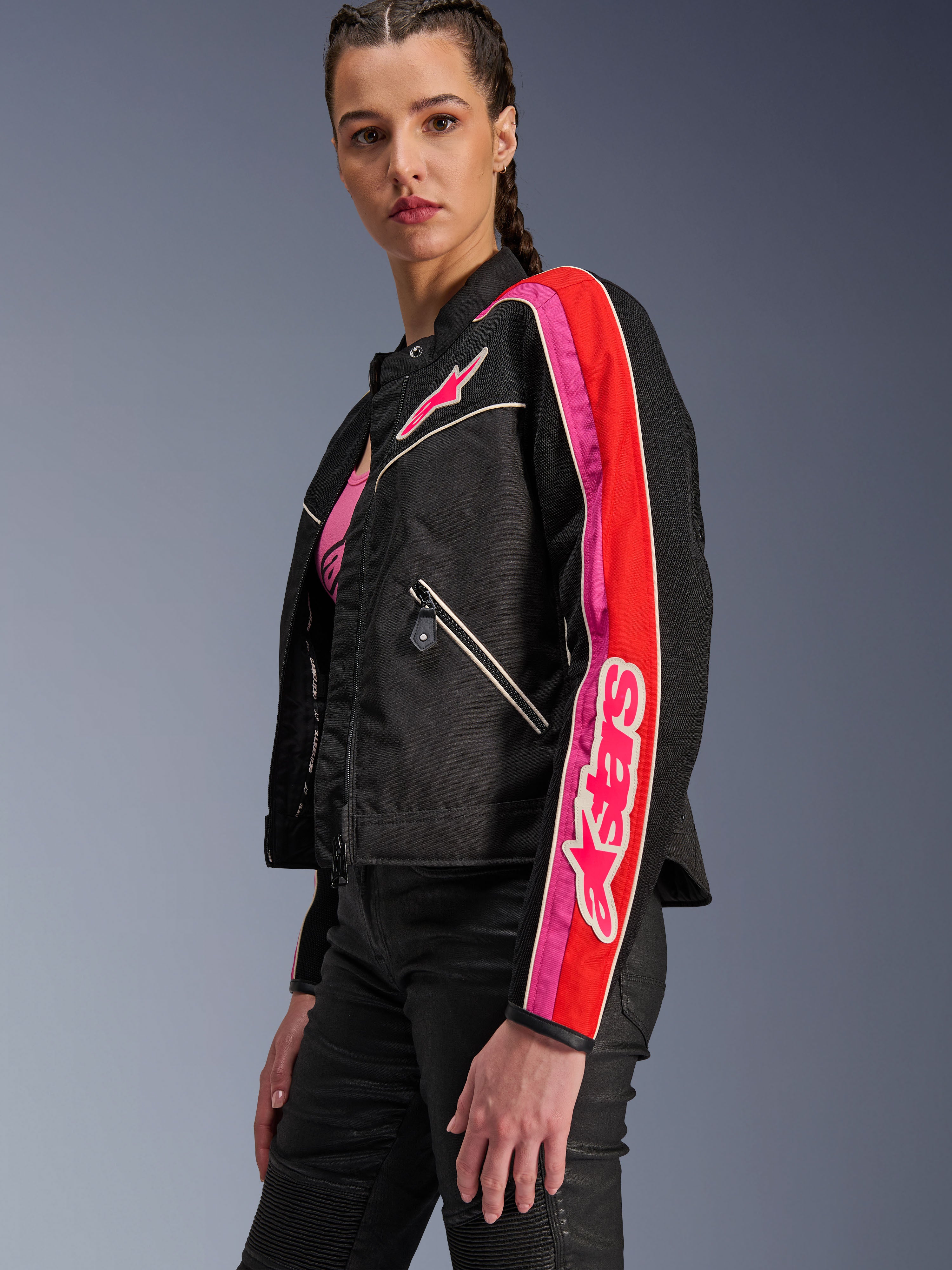 Alpinestars Stella T-Dyno Air Jacket, damska kurtka motocyklowa, czarna z jaskrawoczerwonymi i fuksjowymi akcentami, noszona z czarnymi spodniami, z różowym logo Astars na piersi i rękawie, taliowany motocyklowy krój z panelami z siatki zapewniającymi wentylację
