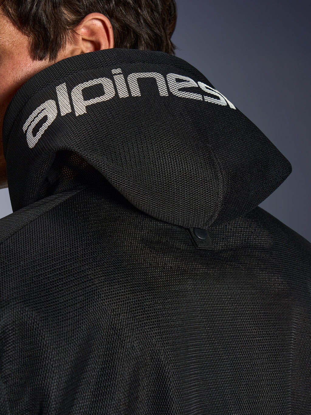 Alpinestars Chrome Superairflow Hoodie, czarna, szczegółowy widok kaptura z siatki 3D z białym brandingiem Alpinestars na górze, widoczny tylny zatrzask stabilizujący, wentylowana tekstylna odzież motocyklowa na ekstremalne upały