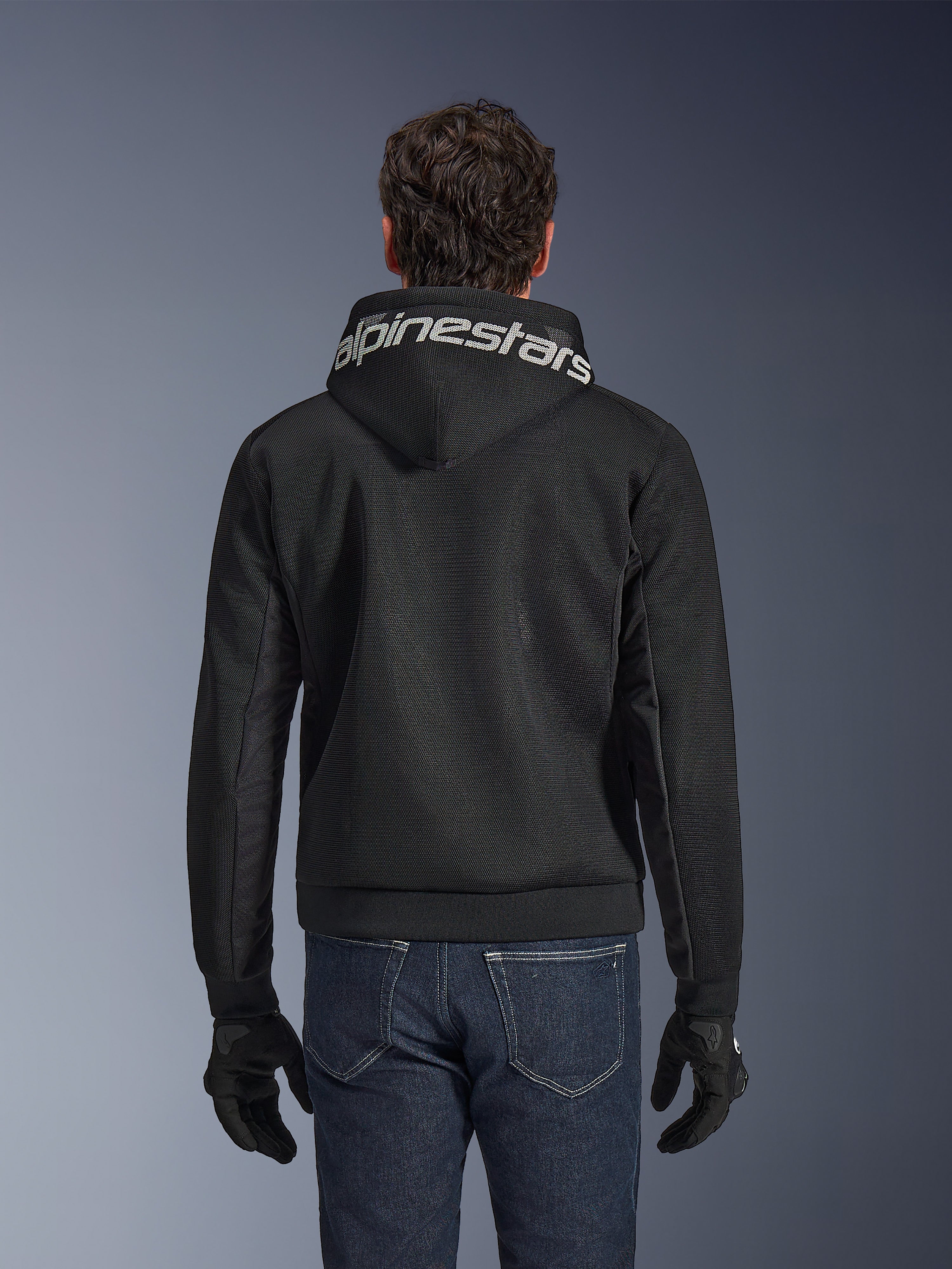 Alpinestars Chrome Superairflow Hoodie, czarna bluza motocyklowa, widok z tyłu z wyrazistym białym logo Alpinestars na kapturze, konstrukcja z technicznej siatki 3D, noszona z ciemnoniebieskimi dżinsami i czarnymi rękawicami, wentylowana odzież do jazdy miejskiej