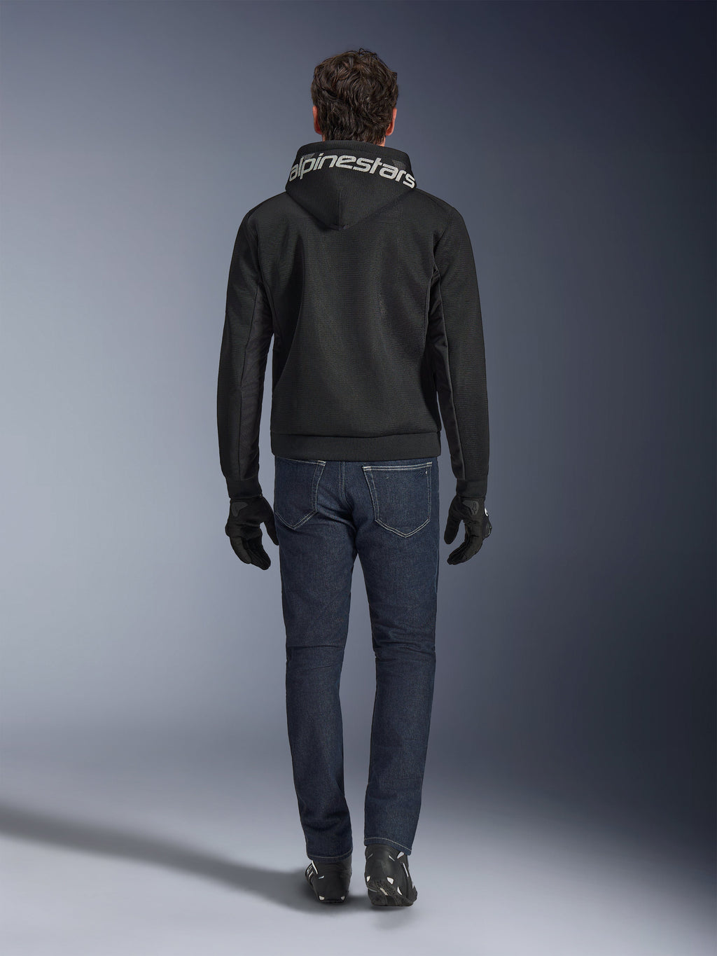Alpinestars Chrome Superairflow Hoodie, czarna bluza motocyklowa z białym logo na kapturze, widok z tyłu, zestawiona z ciemnoniebieskimi dżinsami, czarnymi rękawicami i czarno-białymi butami, miejska sportowa odzież motocyklowa