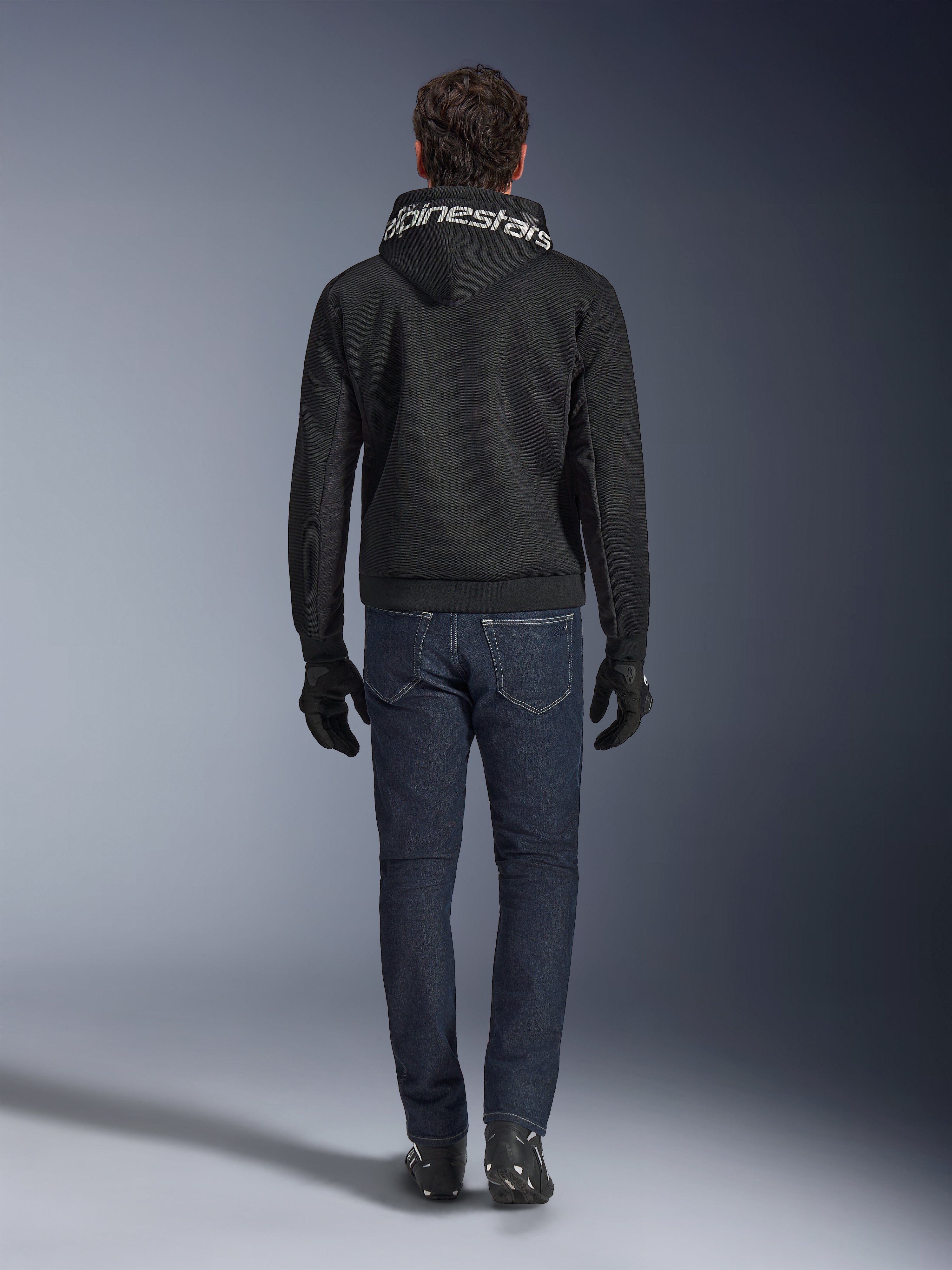 Alpinestars Chrome Superairflow Hoodie, czarna bluza motocyklowa z białym logo na kapturze, widok z tyłu, zestawiona z ciemnoniebieskimi dżinsami, czarnymi rękawicami i czarno-białymi butami, miejska sportowa odzież motocyklowa