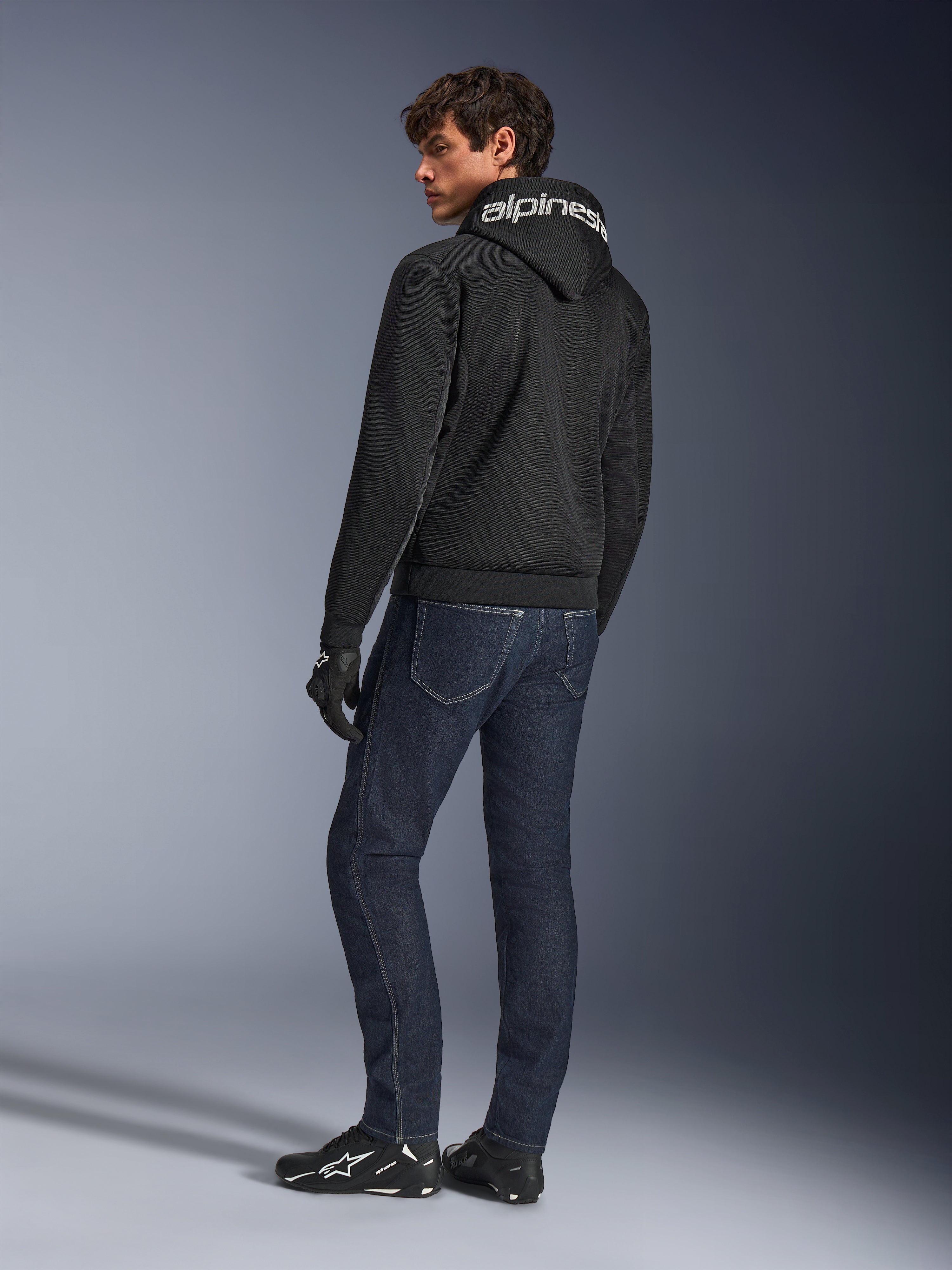 Alpinestars Chrome Superairflow Hoodie, czarna, widok z tyłu na modelu, z białym logo Alpinestars na kapturze, noszona z ciemnymi jeansami i czarnymi rękawicami, miejski styl motocyklowy