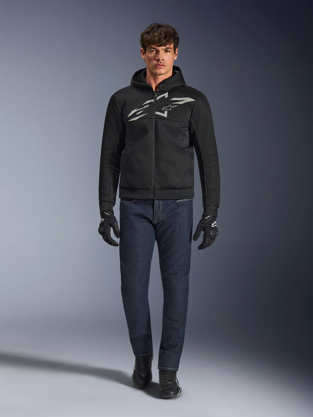 Motocyklista ubrany w bluzę Alpinestars Chrome Superairflow Hoodie w kolorze czarno-szarym, połączoną z ciemnoniebieskimi dżinsami, czarnymi rękawicami motocyklowymi i czarnymi butami. Bluza posiada duże stylizowane logo na piersi oraz strukturę z siatki 3D zapewniającą wydajność w wysokich temperaturach.
