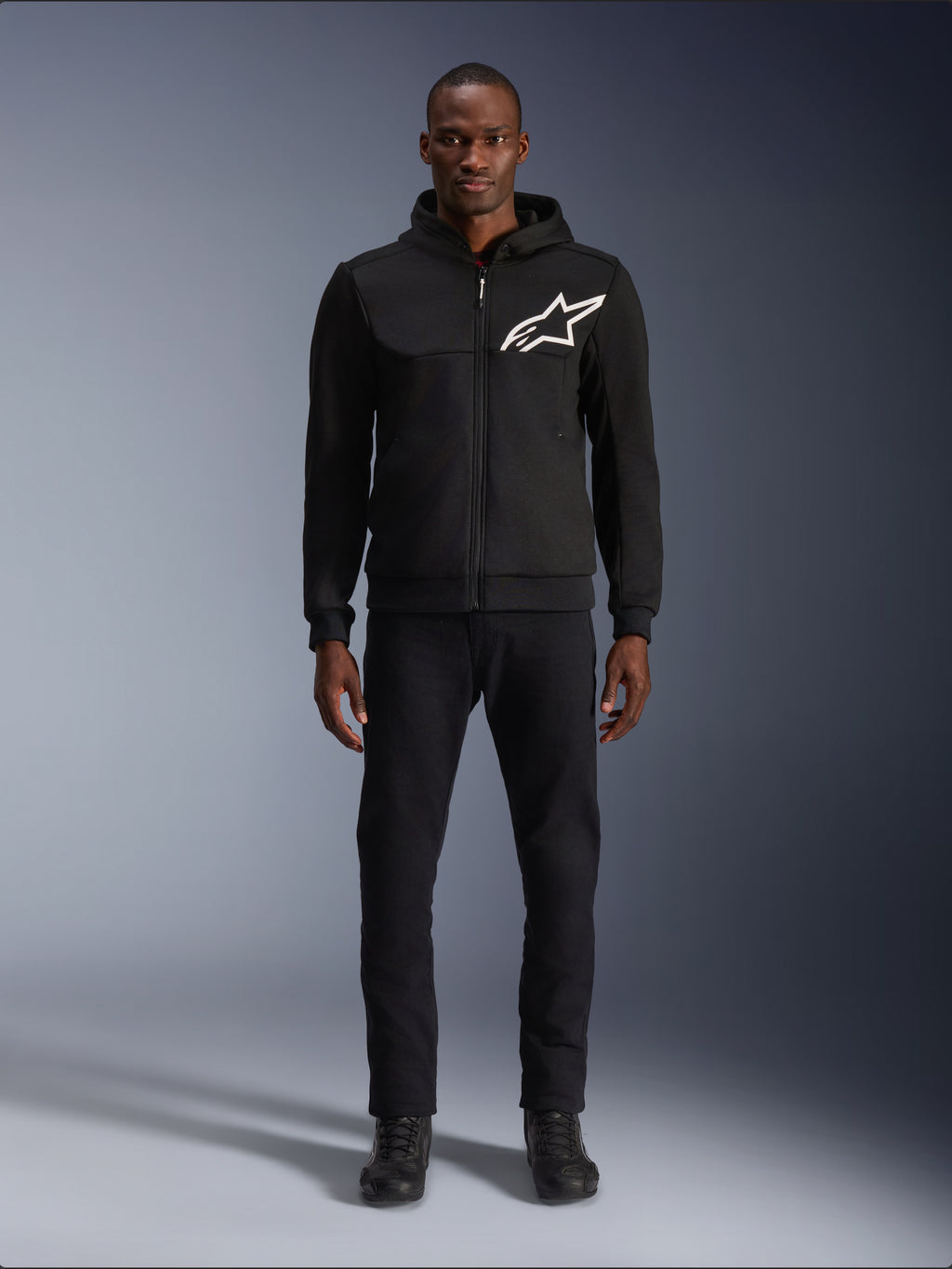 Alpinestars Chrome V2 Sport Hoodie, czarno-biała bluza motocyklowa z dużym białym logo Alpinestars na piersi, zapięcie na zamek i kaptur, prezentowana na modelu w czarnych spodniach i butach, miejska ochronna odzież motocyklowa.