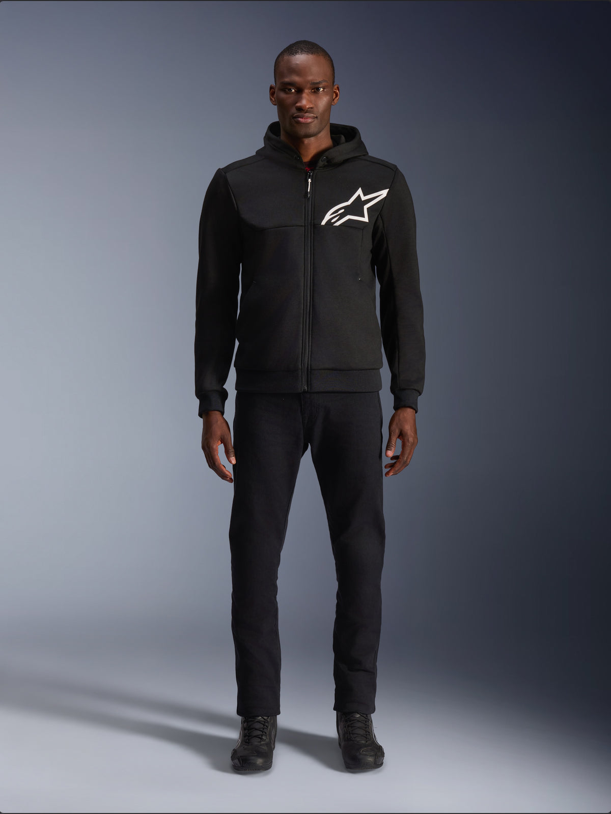 Alpinestars Chrome V2 Sport Hoodie, czarno-biała bluza motocyklowa z dużym białym logo Alpinestars na piersi, zapięcie na zamek i kaptur, prezentowana na modelu w czarnych spodniach i butach, miejska ochronna odzież motocyklowa.