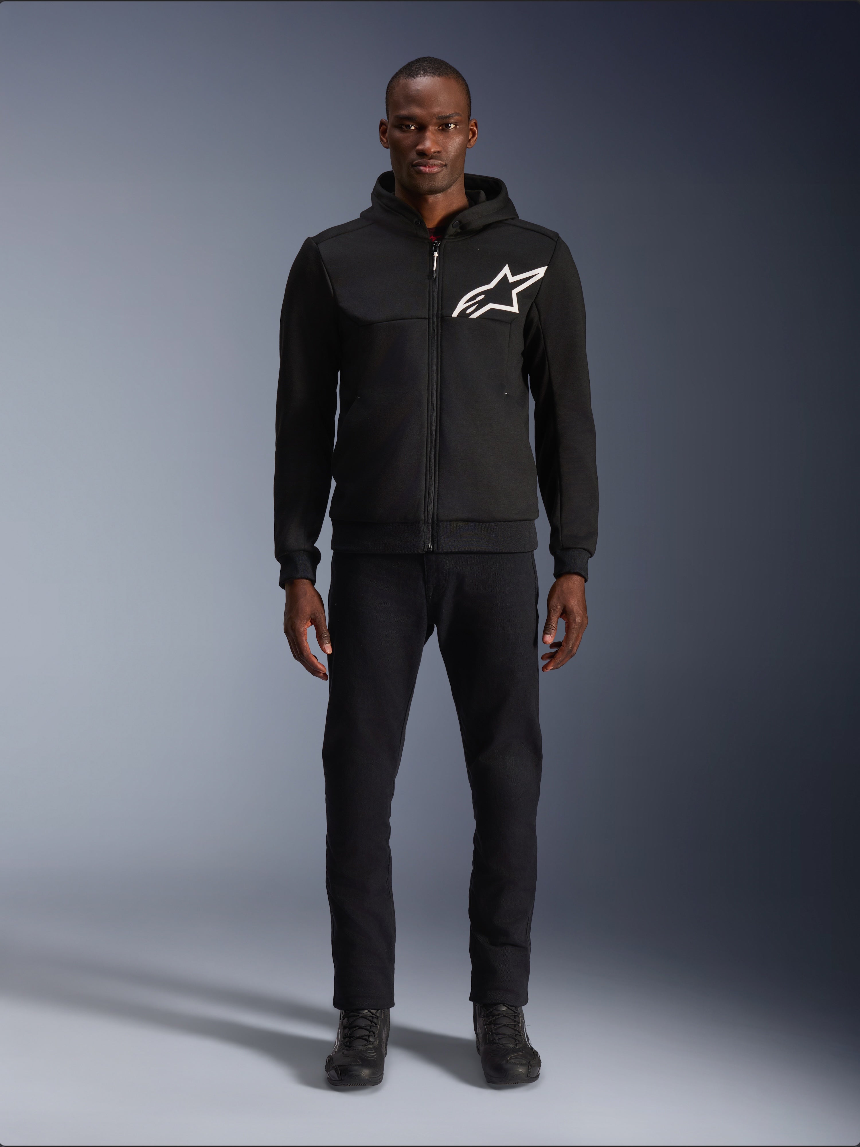Alpinestars Chrome V2 Sport Hoodie, czarno-biała bluza motocyklowa z dużym białym logo Alpinestars na piersi, zapięcie na zamek i kaptur, prezentowana na modelu w czarnych spodniach i butach, miejska ochronna odzież motocyklowa.
