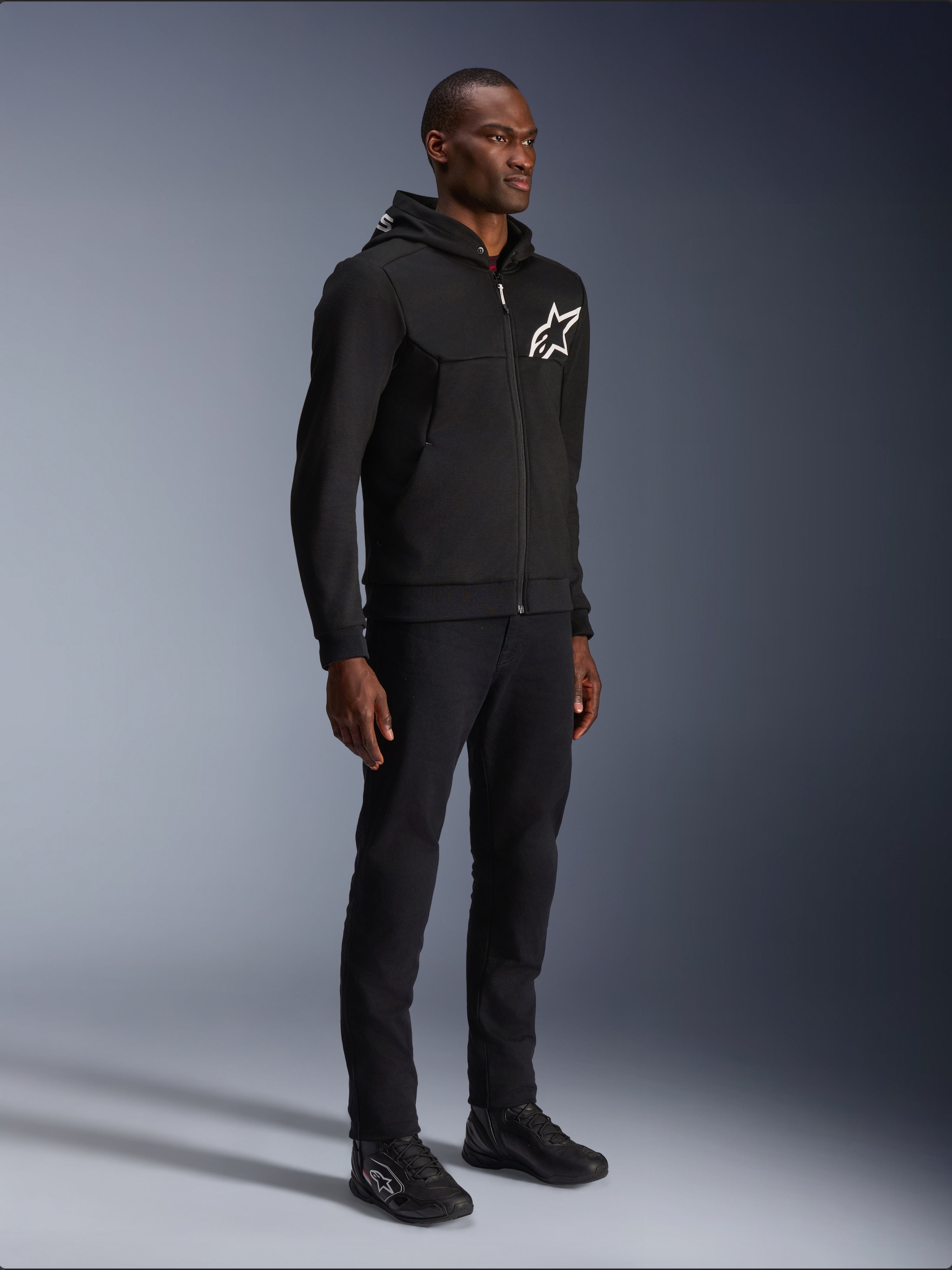 Alpinestars Chrome V2 Sport Hoodie, czarna bluza motocyklowa z białym logo gwiazdy, prezentowana przez modela w pasujących czarnych spodniach i butach motocyklowych typu sneakers, miejska odzież motocyklowa lifestyle