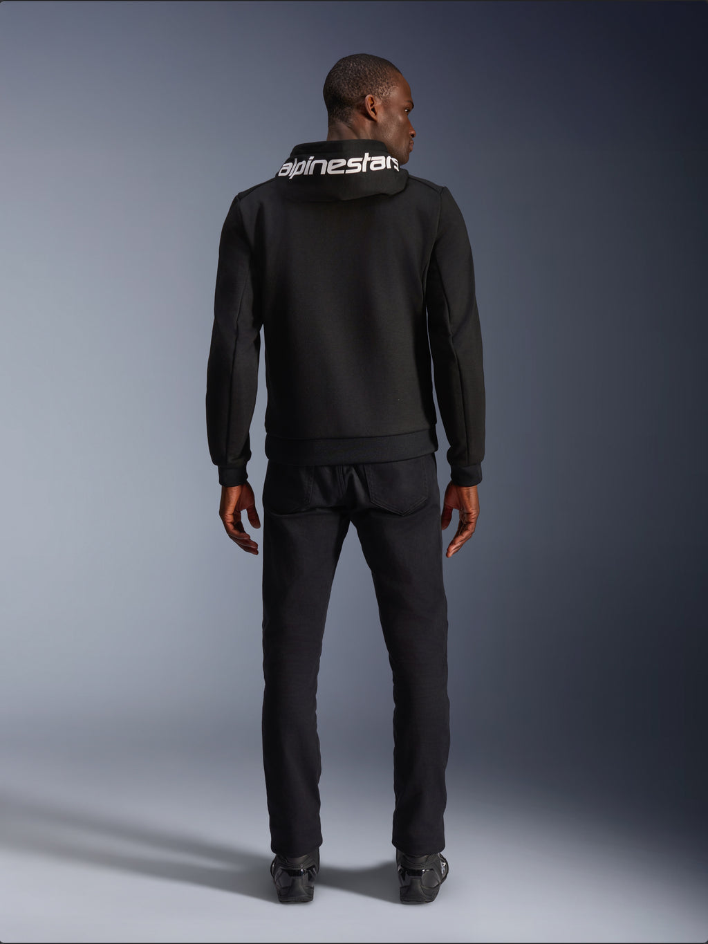 Alpinestars Chrome V2 Sport Hoodie, motocyklowa bluza z kapturem, czarna, widok z tyłu modelu ubranego w czarną bluzę z polaru tech z białym logo Alpinestars na kapturze, w zestawieniu z czarnymi spodniami i butami, casualowy miejski styl jazdy