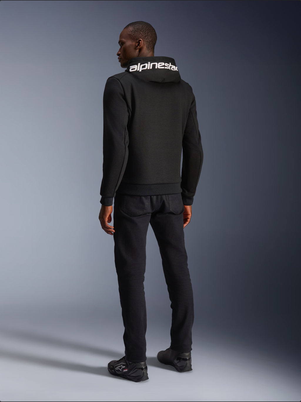 Alpinestars Chrome V2 Sport Hoodie, czarna, widok z tyłu prezentujący biały branding Alpinestars na kapturze, noszona z czarnymi spodniami i butami motocyklowymi, miejska odzież motocyklowa o sportowej sylwetce