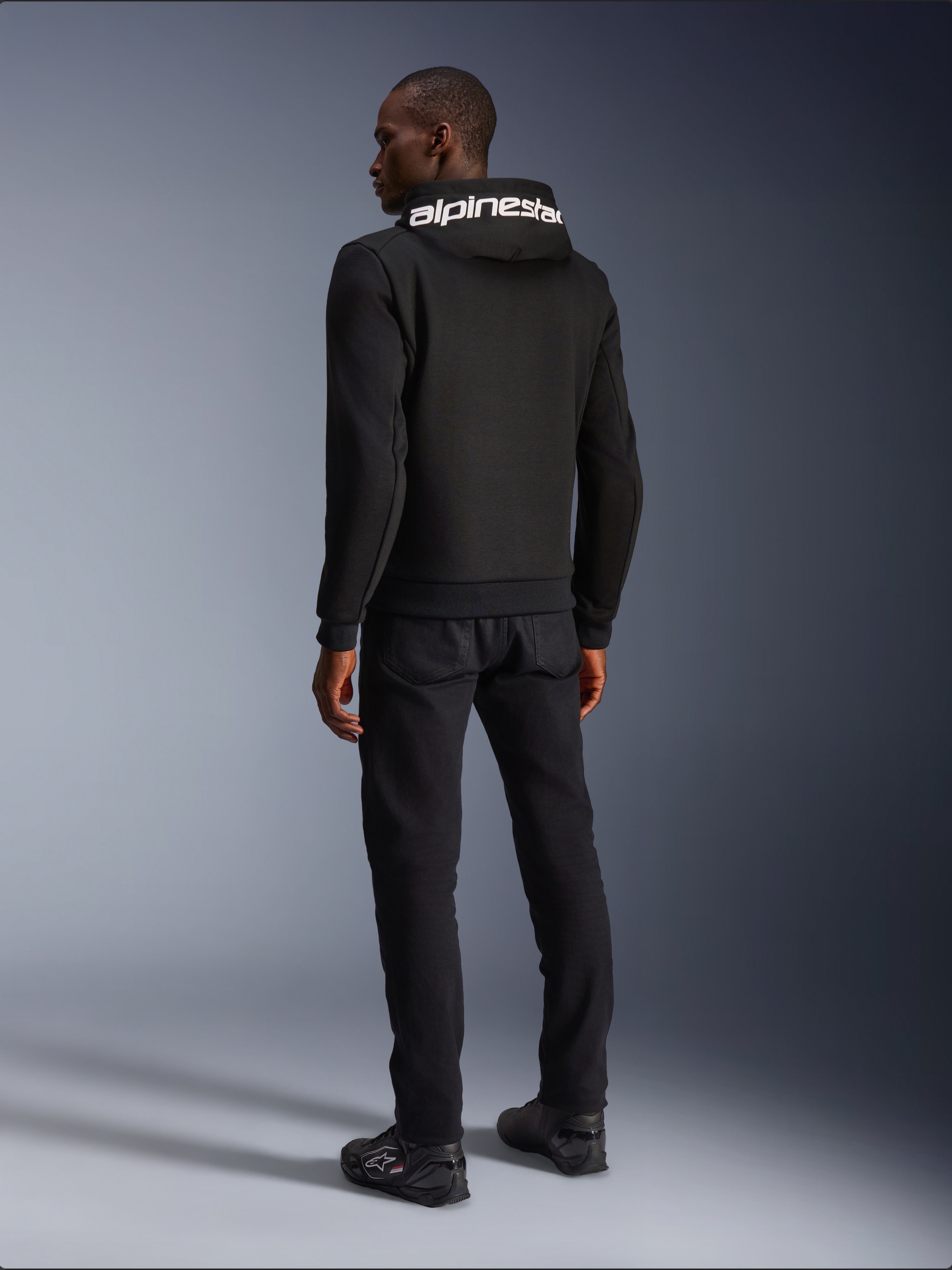 Alpinestars Chrome V2 Sport Hoodie, czarna, widok z tyłu prezentujący biały branding Alpinestars na kapturze, noszona z czarnymi spodniami i butami motocyklowymi, miejska odzież motocyklowa o sportowej sylwetce