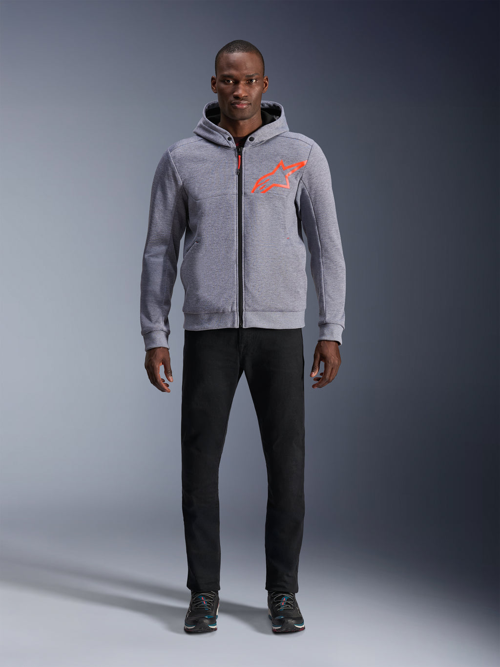 Model w bluzie z kapturem Alpinestars Chrome V2 Sport Hoodie w kolorze Gray Heather Red Fluo, jasnoszara techniczna bluza z polaru z jaskrawym pomarańczowo-czerwonym logo gwiazdy na piersi, czarne spodnie i ciemne sneakersy, casualowy miejski styl motocyklowy