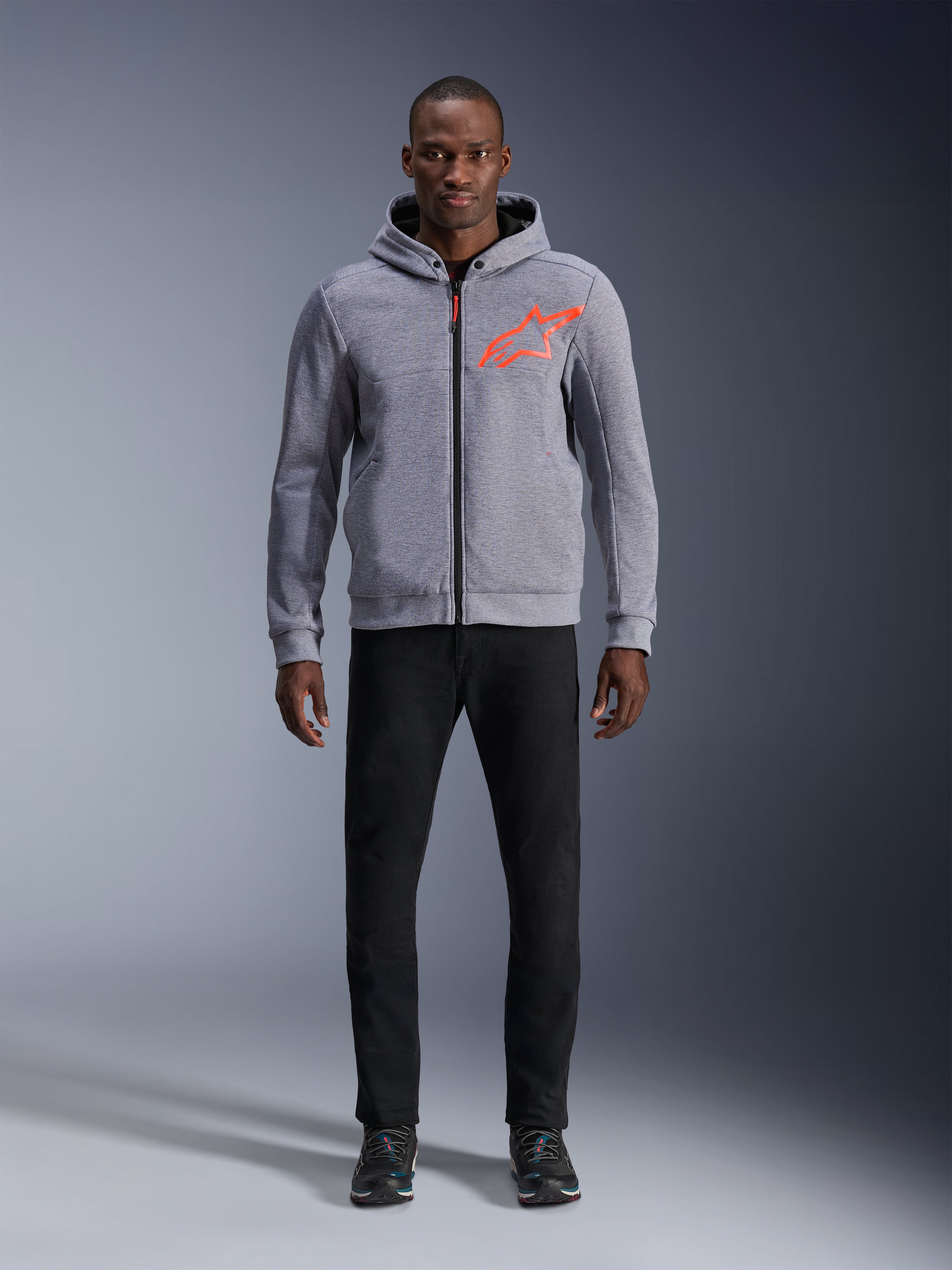 Model w bluzie z kapturem Alpinestars Chrome V2 Sport Hoodie w kolorze Gray Heather Red Fluo, jasnoszara techniczna bluza z polaru z jaskrawym pomarańczowo-czerwonym logo gwiazdy na piersi, czarne spodnie i ciemne sneakersy, casualowy miejski styl motocyklowy