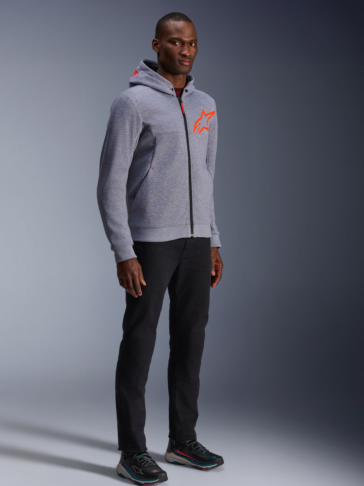 Alpinestars Chrome V2 Sport Hoodie, szary melanż i czerwony fluo, noszona przez modela w czarnych spodniach i ciemnych butach sportowych, z jaskrawopomarańczowym logo gwiazdy na piersi i kapturze, codzienna miejska odzież motocyklowa