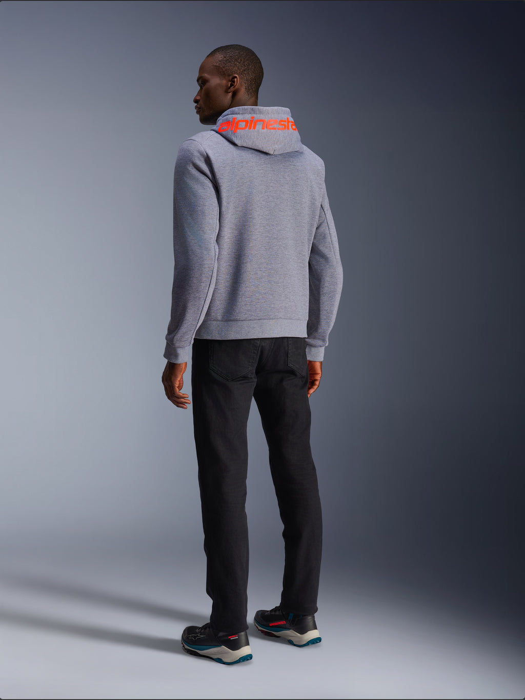 Alpinestars Chrome V2 Sport Hoodie, bluza z kapturem w kolorze szarym melanżowym, widok z tyłu z dużym fluorescencyjnym czerwonym napisem Alpinestars na kapturze, model w czarnych spodniach i ciemnych butach motocyklowych, codzienna miejska odzież motocyklowa