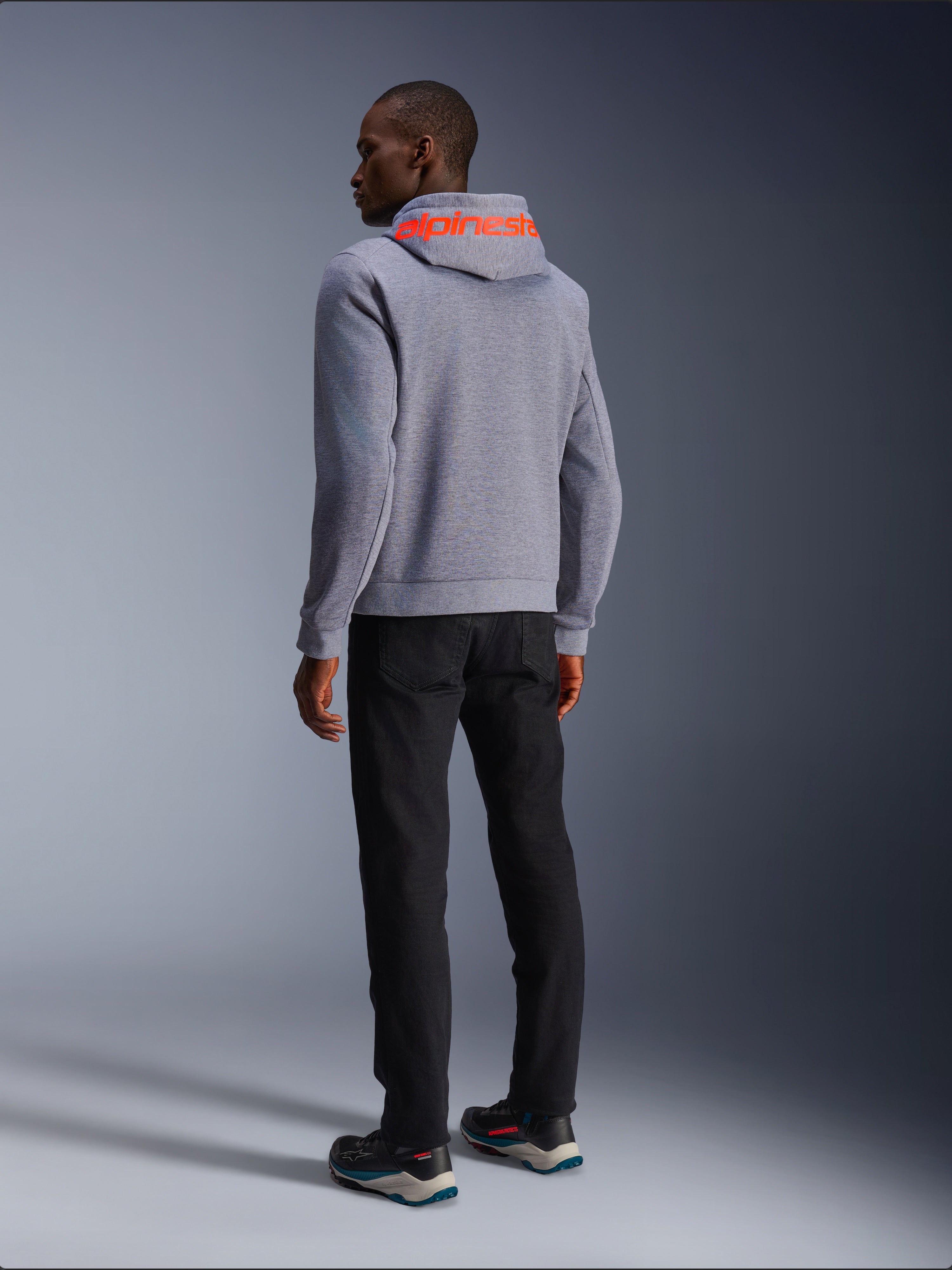 Alpinestars Chrome V2 Sport Hoodie, bluza z kapturem w kolorze szarym melanżowym, widok z tyłu z dużym fluorescencyjnym czerwonym napisem Alpinestars na kapturze, model w czarnych spodniach i ciemnych butach motocyklowych, codzienna miejska odzież motocyklowa