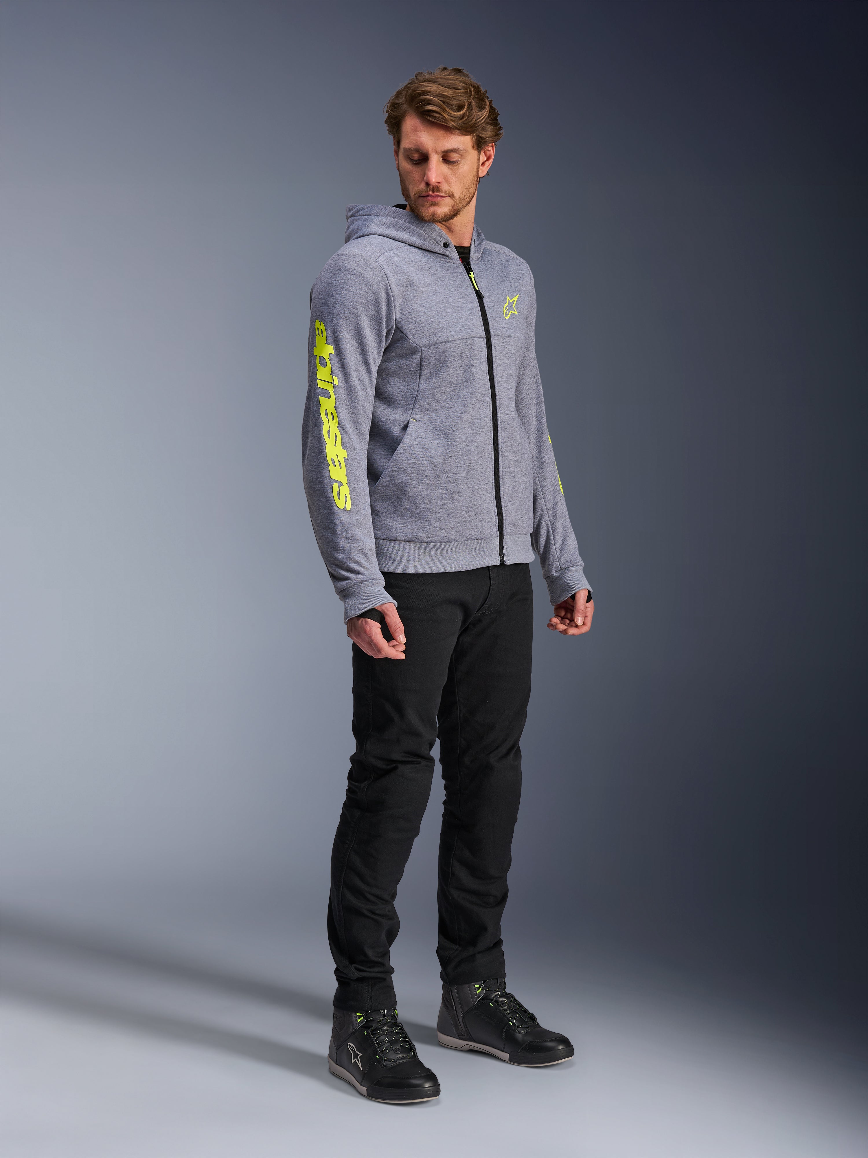 Alpinestars Chrome V2 Sport Hoodie, bluza motocyklowa z kapturem, szary melanż i neonowy żółty, noszona przez modela w czarnych spodniach i butach motocyklowych, wyraziste żółte logo na rękawie i klatce piersiowej, miejska odzież motocyklowa