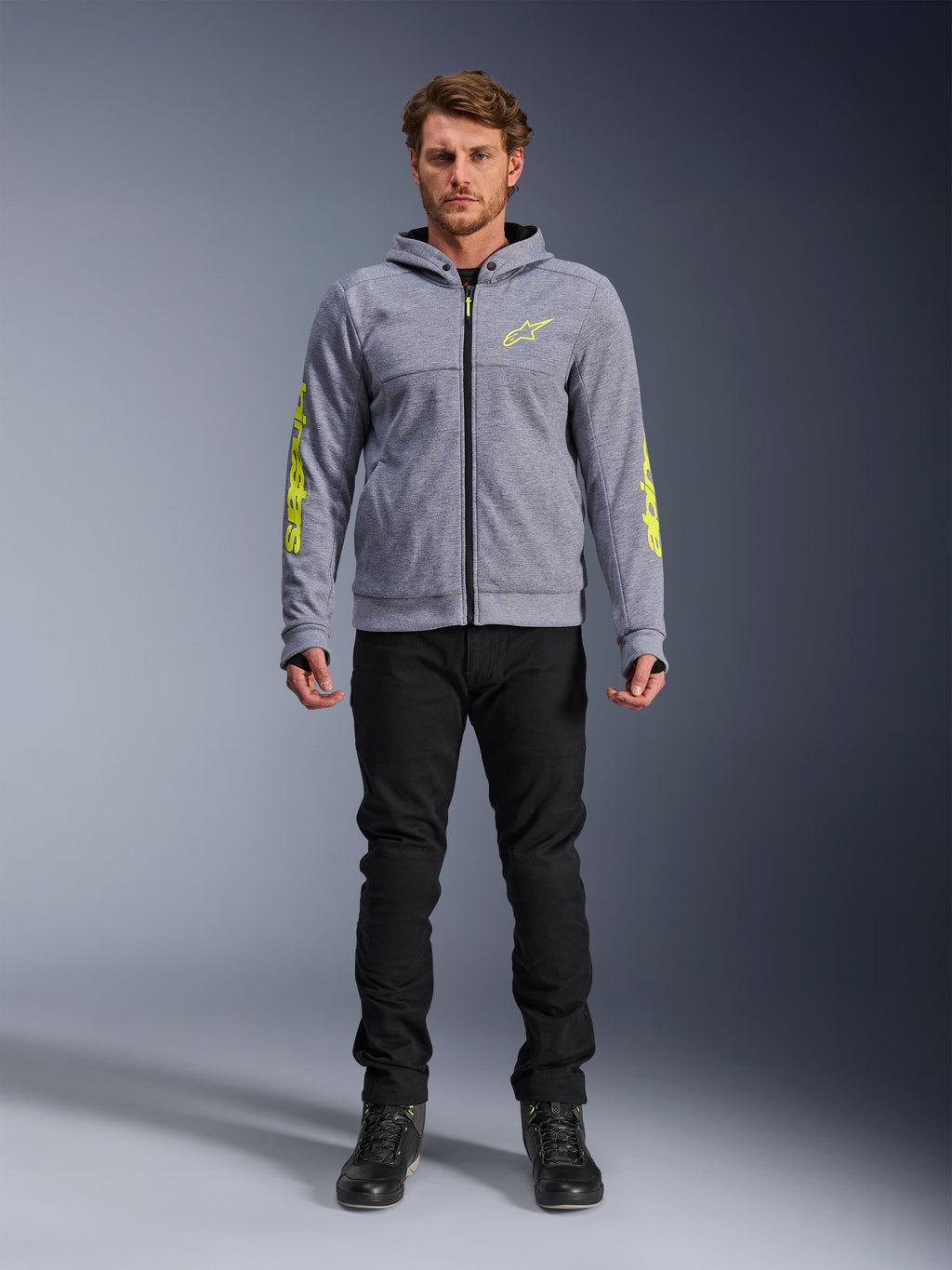 Bluza motocyklowa Alpinestars Chrome V2 Sport Hoodie, szary melanż z jaskrawożółtymi akcentami, miejska odzież motocyklowa, zestawiona z czarnymi spodniami i czarnymi butami motocyklowymi, techniczny polar z wyrazistym żółtym logo na rękawach i klatce piersiowej, swobodny styl miejski