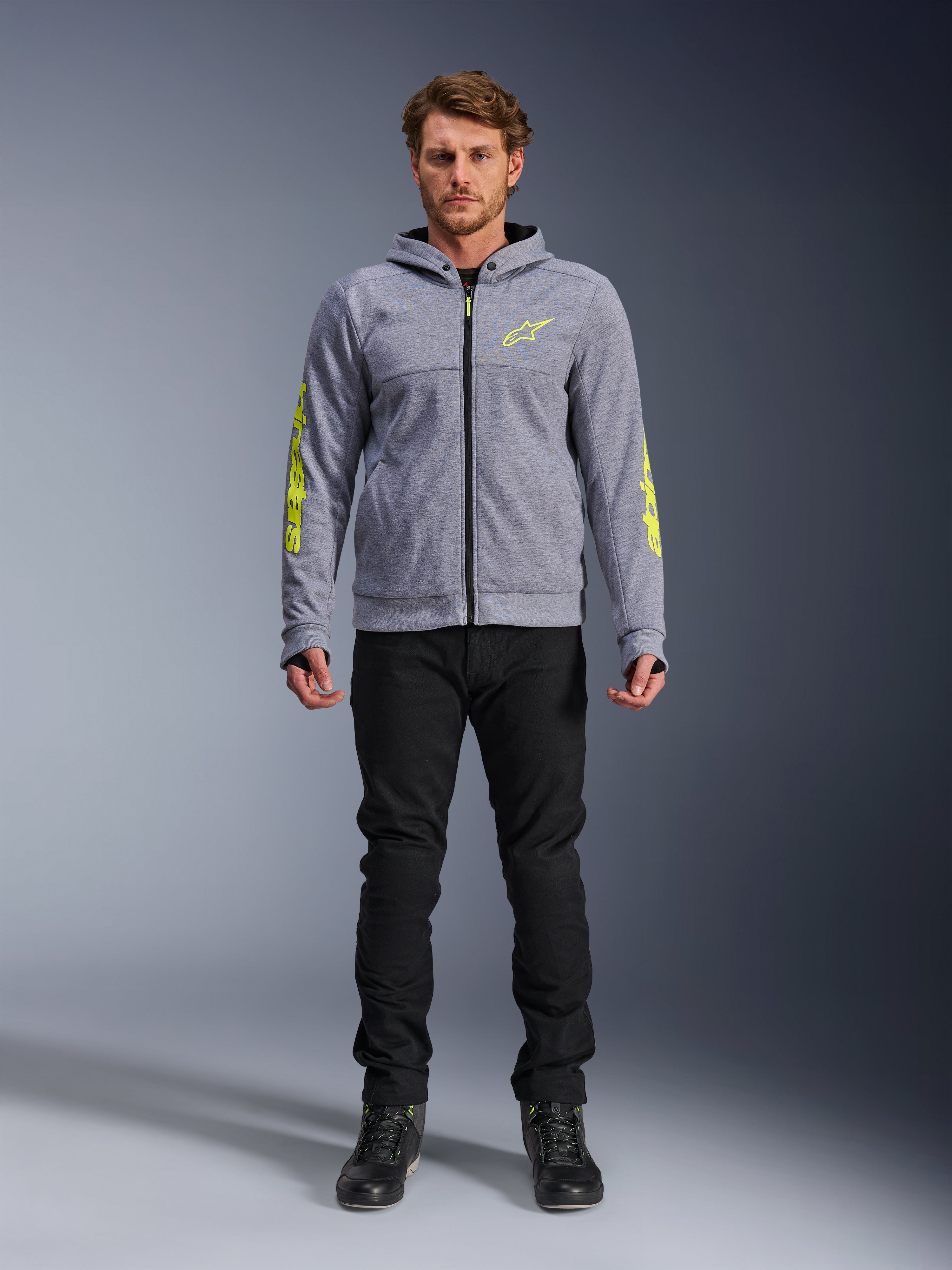 Bluza motocyklowa Alpinestars Chrome V2 Sport Hoodie, szary melanż z jaskrawożółtymi akcentami, miejska odzież motocyklowa, zestawiona z czarnymi spodniami i czarnymi butami motocyklowymi, techniczny polar z wyrazistym żółtym logo na rękawach i klatce piersiowej, swobodny styl miejski