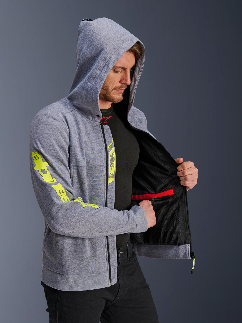 Alpinestars Chrome V2 Sport Hoodie, szary melanż i fluorescencyjny żółty, mężczyzna w bluzie z założonym kapturem, rozpięta ukazująca czarną siatkową podszewkę i wewnętrzną czerwoną kieszeń na zamek, duże żółte logo na rękawie, codzienna miejska odzież motocyklowa