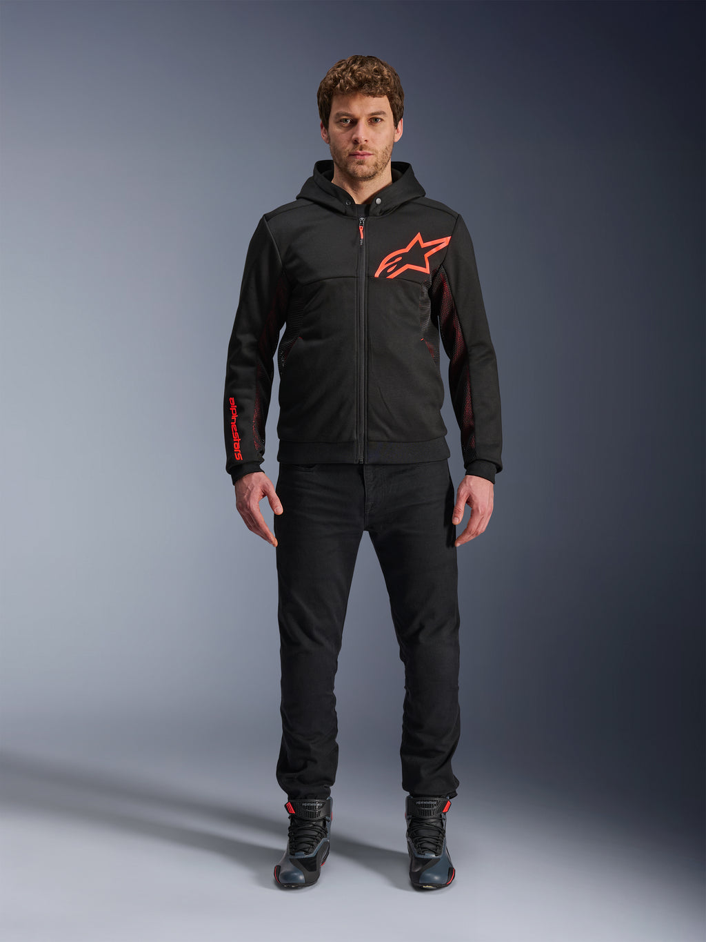 Alpinestars Chrome Air Sport Hoodie, czarno-czerwona fluo, miejska kurtka motocyklowa o konstrukcji z polaru tech, z dużym czerwonym logo Astars na klatce piersiowej i brandingiem na rękawie, noszona przez modela z czarnymi spodniami i butami motocyklowymi