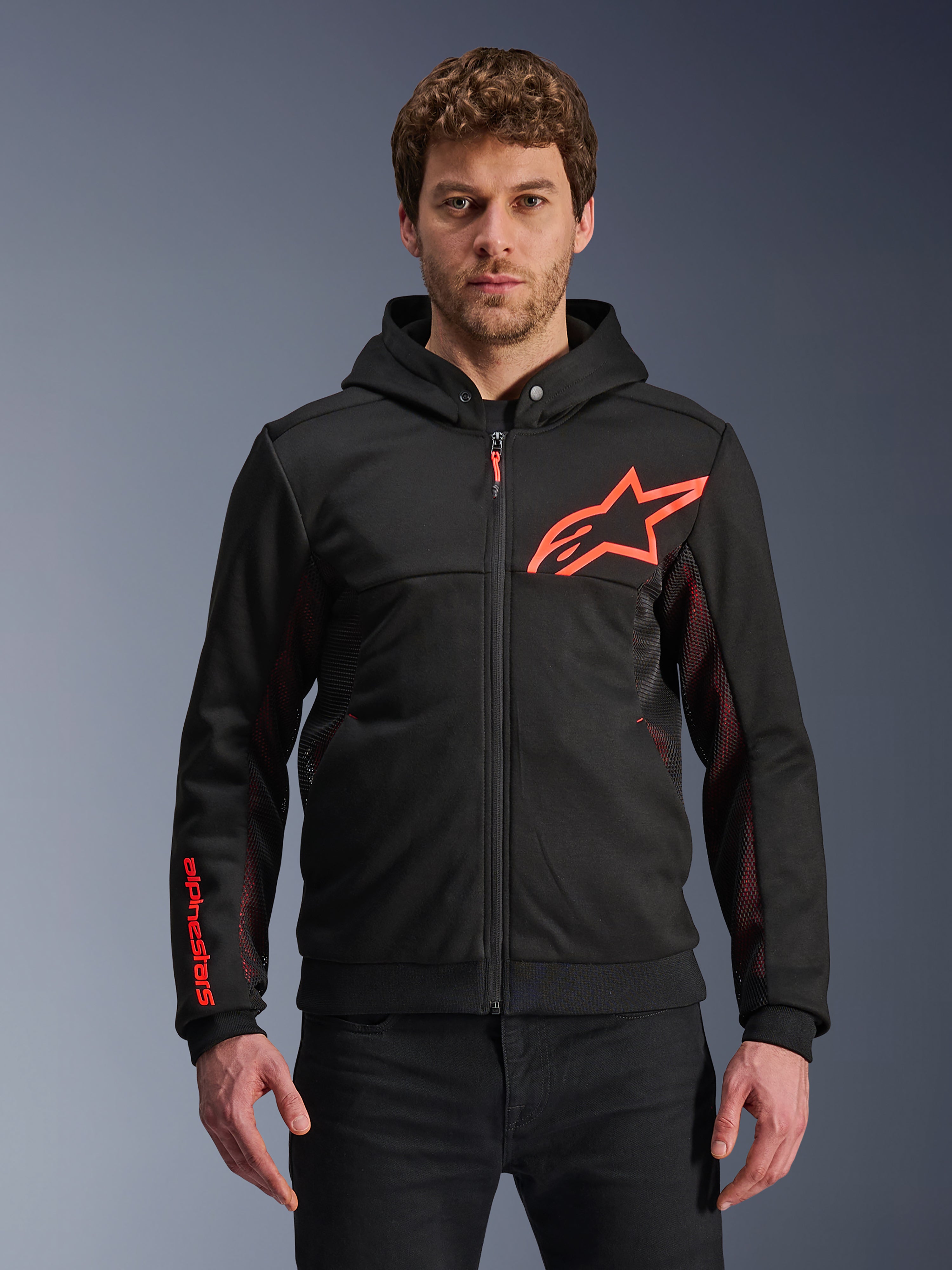 Alpinestars Chrome Air Sport Hoodie, czarna bluza z kapturem na zamek z czerwonym fluorescencyjnym logo gwiazdy i akcentami na rękawach, czerwone panele boczne z siateczki dla wentylacji, miejska odzież motocyklowa casualowa noszona przez modela