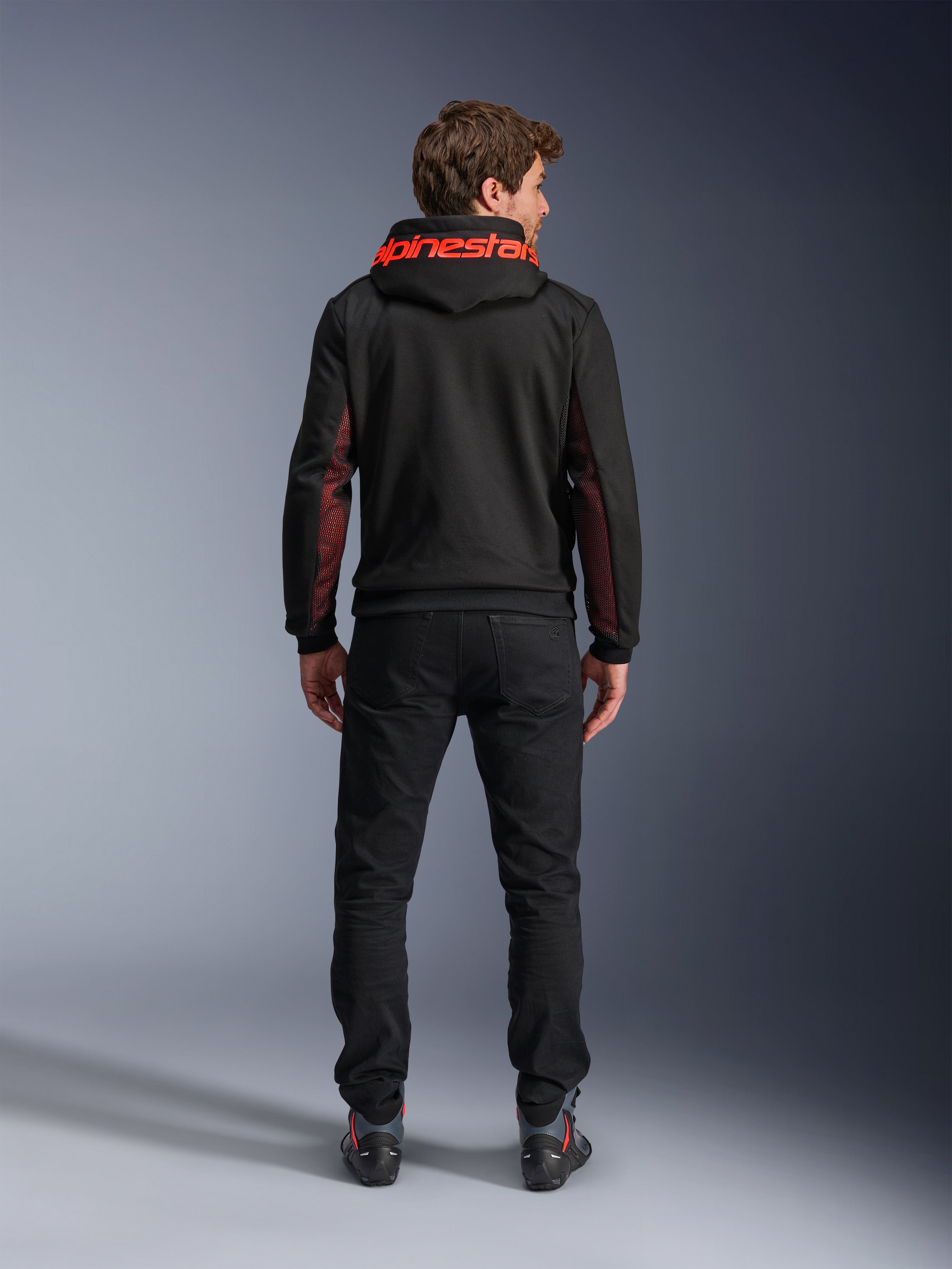Alpinestars Chrome Air Sport Hoodie, czarno-czerwona (Black Red Fluo), widok z tyłu na mężczyznę w miejskiej kurtce motocyklowej, czerwony napis Alpinestars na kapturze, czerwone panele z siatki na rękawach, czarne spodnie