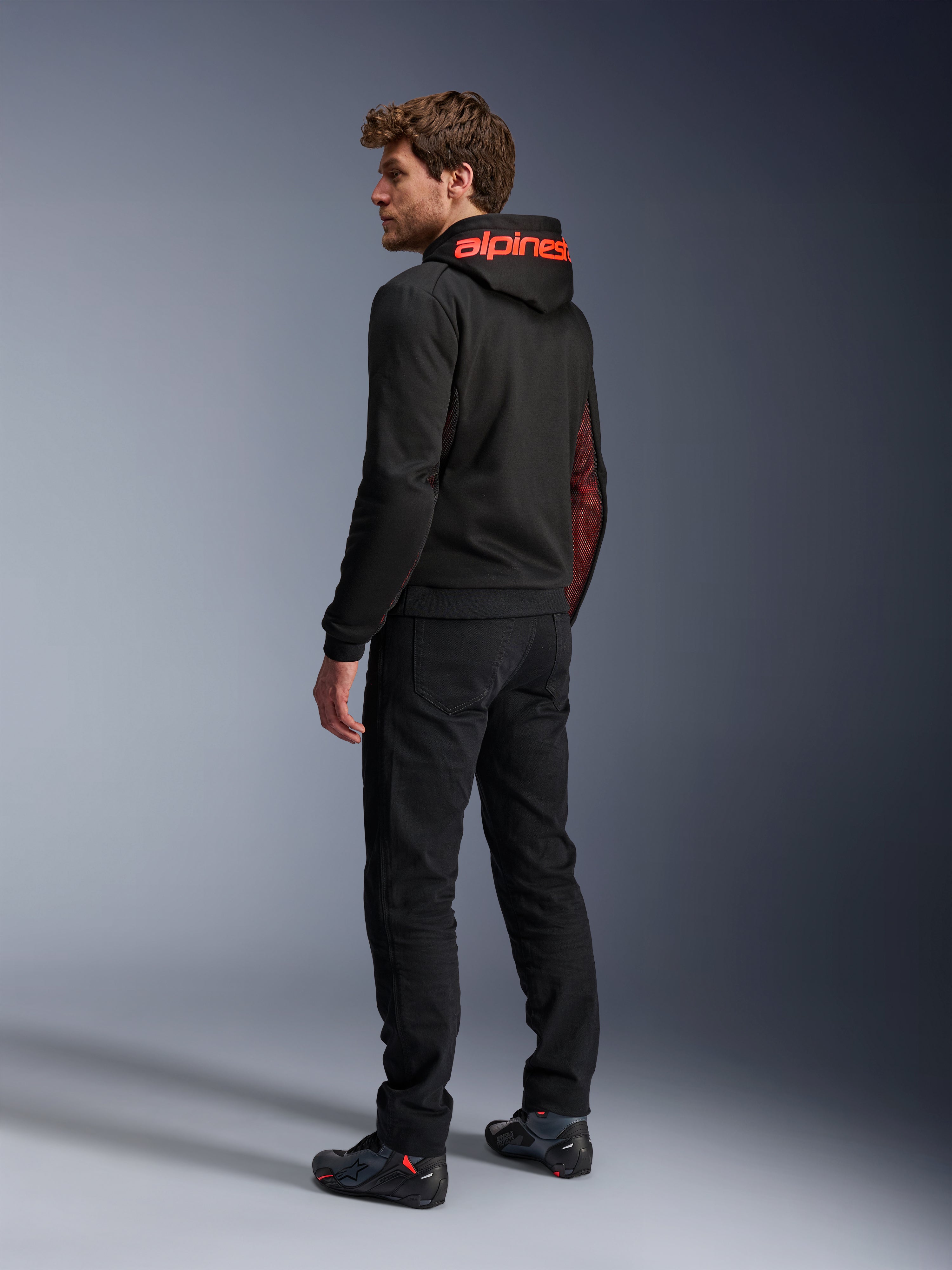 Alpinestars Chrome Air Sport Hoodie, miejska bluza motocyklowa, czarna i czerwona Fluo, widok z tyłu przedstawiający czerwone logo Alpinestars na kapturze i siatkowe panele boczne, noszona z czarnymi spodniami motocyklowymi i obuwiem technicznym