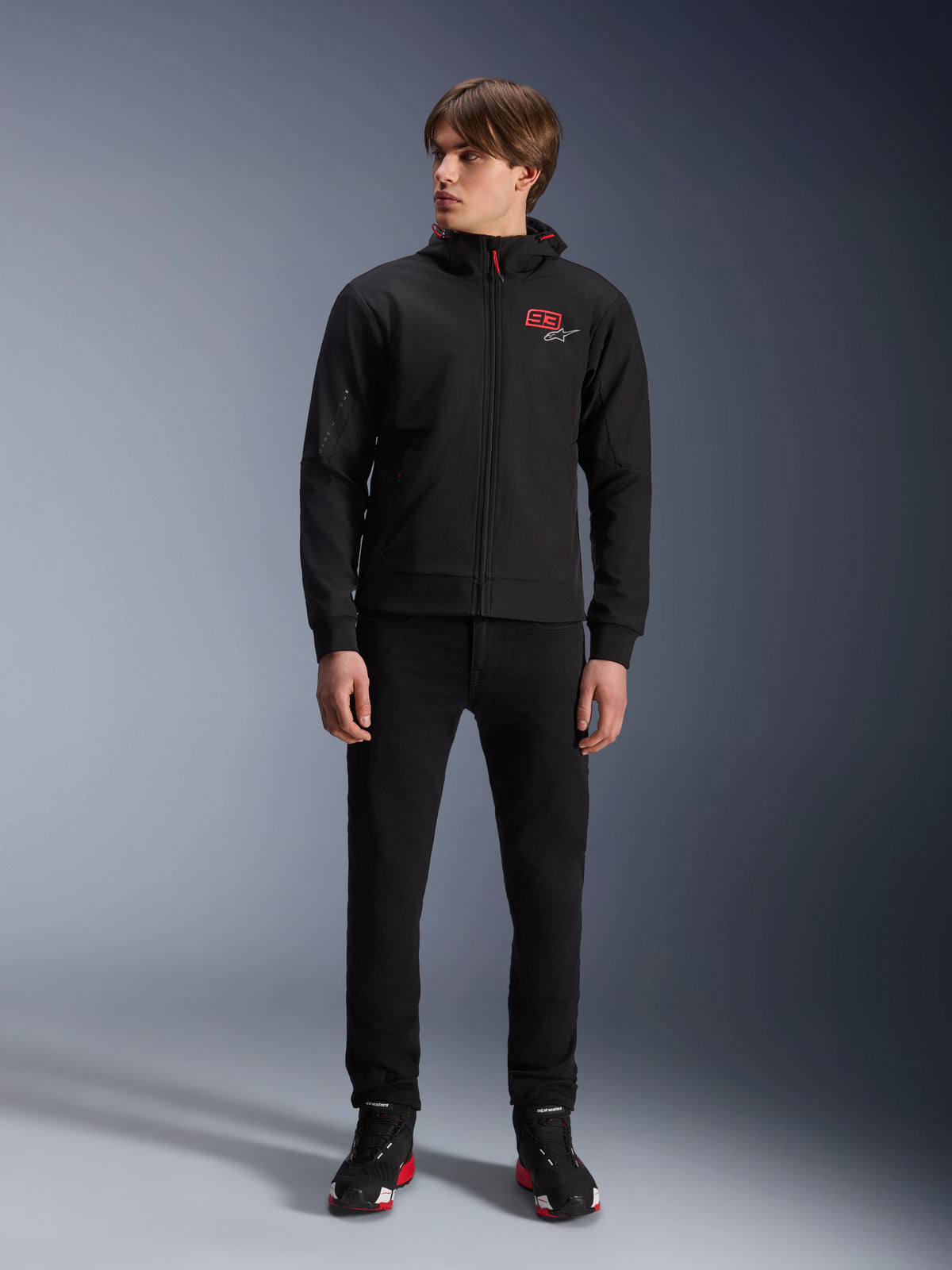 Alpinestars MM93 Radium Tech Hoodie, czarna miejska bluza motocyklowa z kapturem, z czerwonym logo 93 i białym logo gwiazdy na piersi, noszona przez modela z czarnymi spodniami i dopasowanymi butami motocyklowymi, wiatroodporna powłoka techniczna do jazdy miejskiej