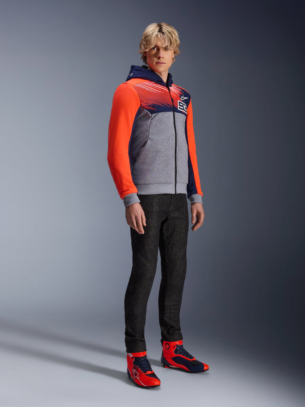 Alpinestars MM93 Chrome V2 Sport Hoodie, miejska bluza motocyklowa, korpus w kolorze szary melanż z granatową klatką piersiową i jaskrawoczerwonymi rękawami fluo, logo 93 na piersi, prezentowana przez modela w czarnych dopasowanych spodniach i pasujących czerwono-niebieskich wysokich butach motocyklowych, nowoczesny sportowy krój do jazdy miejskiej