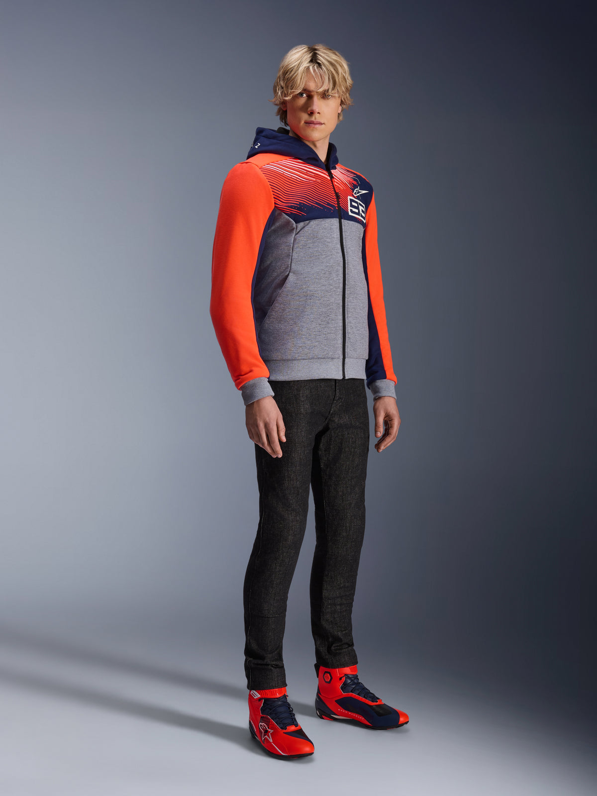 Alpinestars MM93 Chrome V2 Sport Hoodie, miejska bluza motocyklowa, korpus w kolorze szary melanż z granatową klatką piersiową i jaskrawoczerwonymi rękawami fluo, logo 93 na piersi, prezentowana przez modela w czarnych dopasowanych spodniach i pasujących czerwono-niebieskich wysokich butach motocyklowych, nowoczesny sportowy krój do jazdy miejskiej