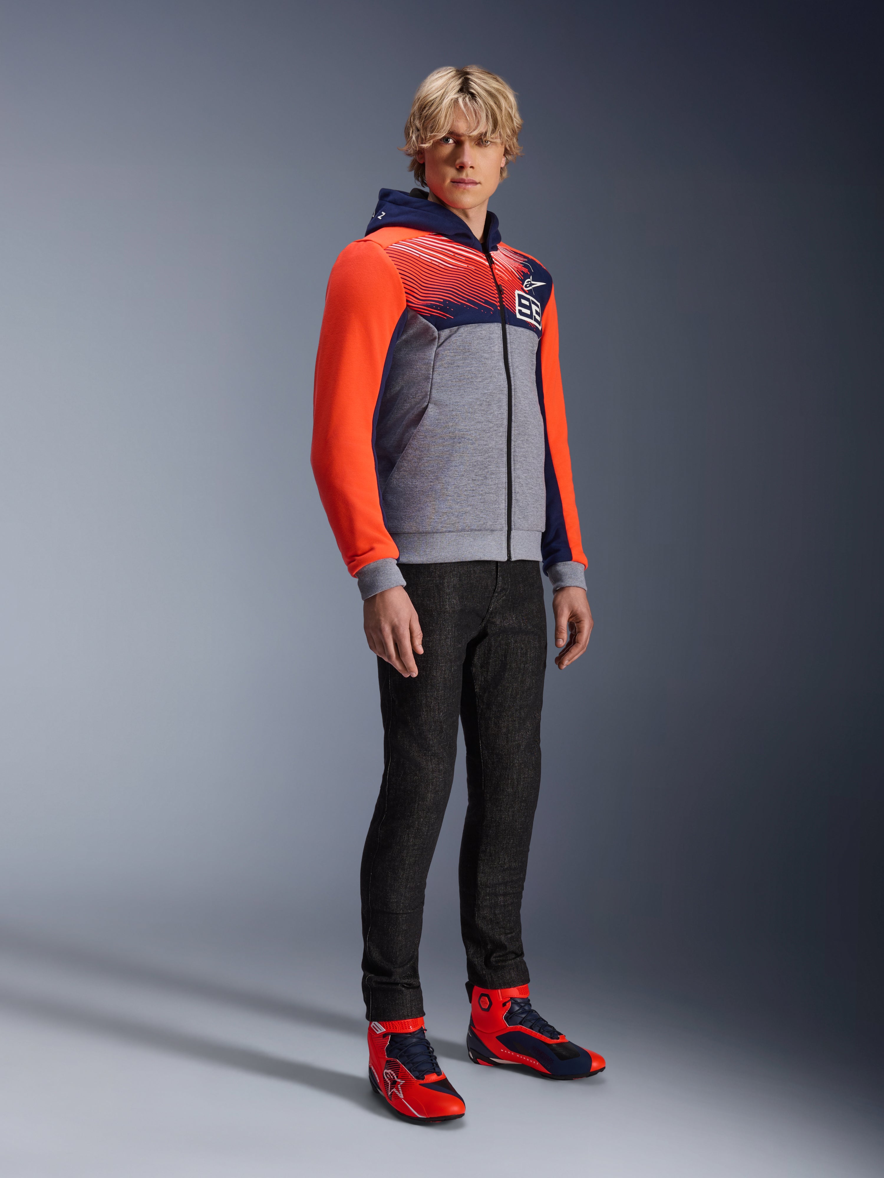 Alpinestars MM93 Chrome V2 Sport Hoodie, miejska bluza motocyklowa, korpus w kolorze szary melanż z granatową klatką piersiową i jaskrawoczerwonymi rękawami fluo, logo 93 na piersi, prezentowana przez modela w czarnych dopasowanych spodniach i pasujących czerwono-niebieskich wysokich butach motocyklowych, nowoczesny sportowy krój do jazdy miejskiej