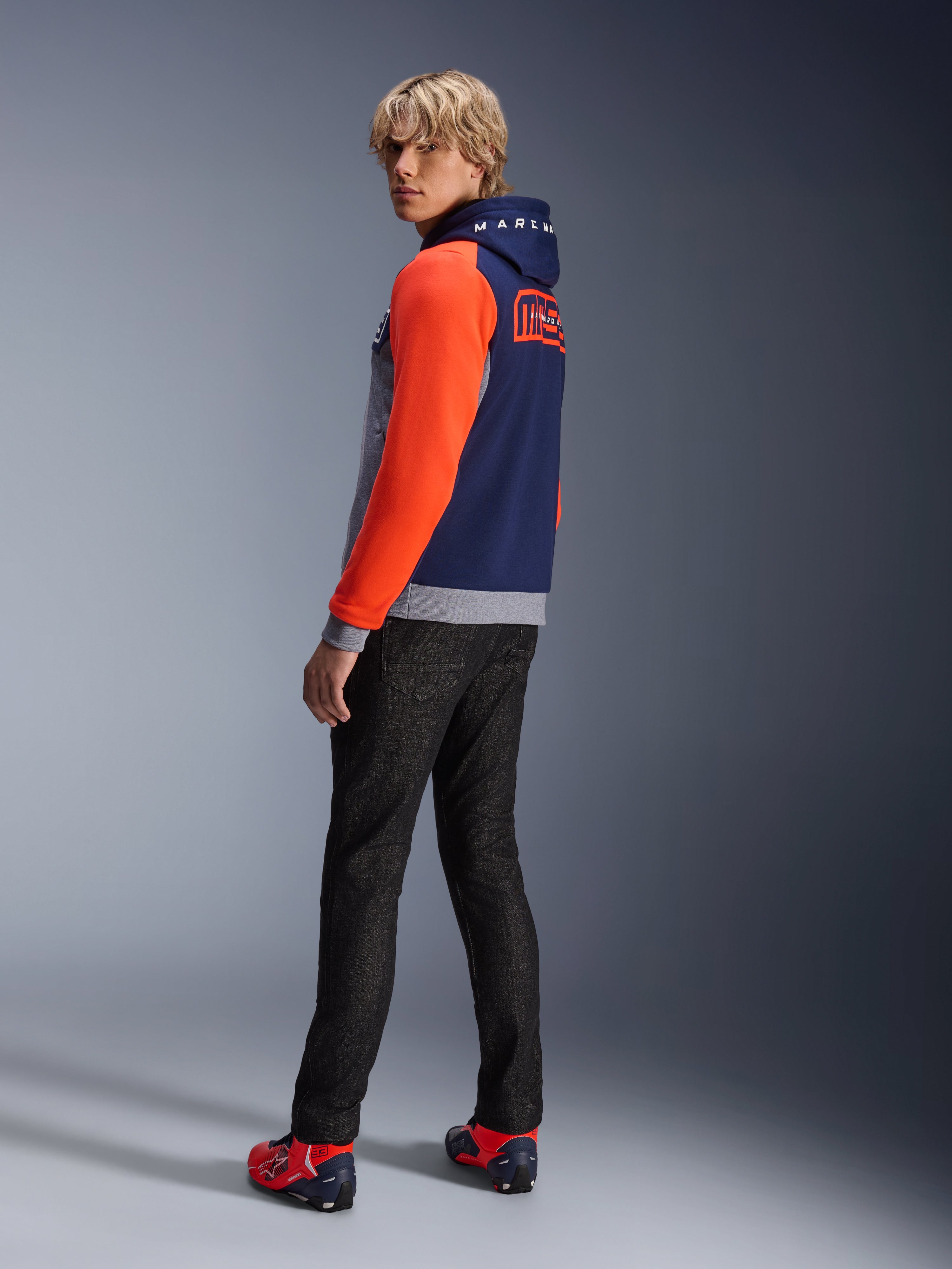 Alpinestars MM93 Chrome V2 Sport Hoodie, granatowo-szara bluza z kapturem z jaskrawoczerwonymi rękawami, branding MM93 Marc Marquez na plecach, model w ciemnych dżinsach i czerwonych butach motocyklowych, codzienna miejska odzież motocyklowa
